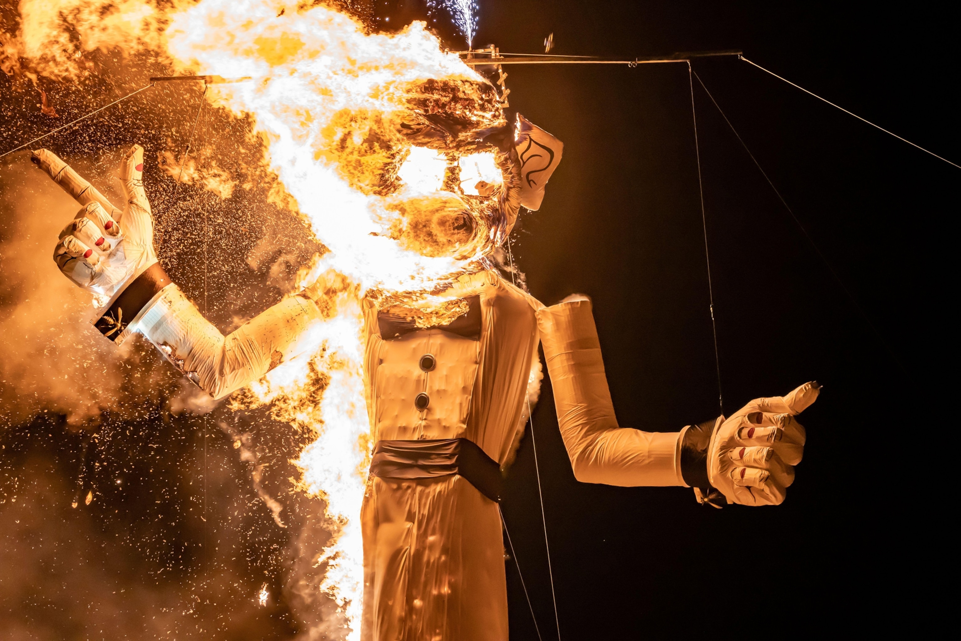 The effigy figure, Zozobra is seen ablaze in flames. It is nighttime and there is an orange glow amongst the burning.