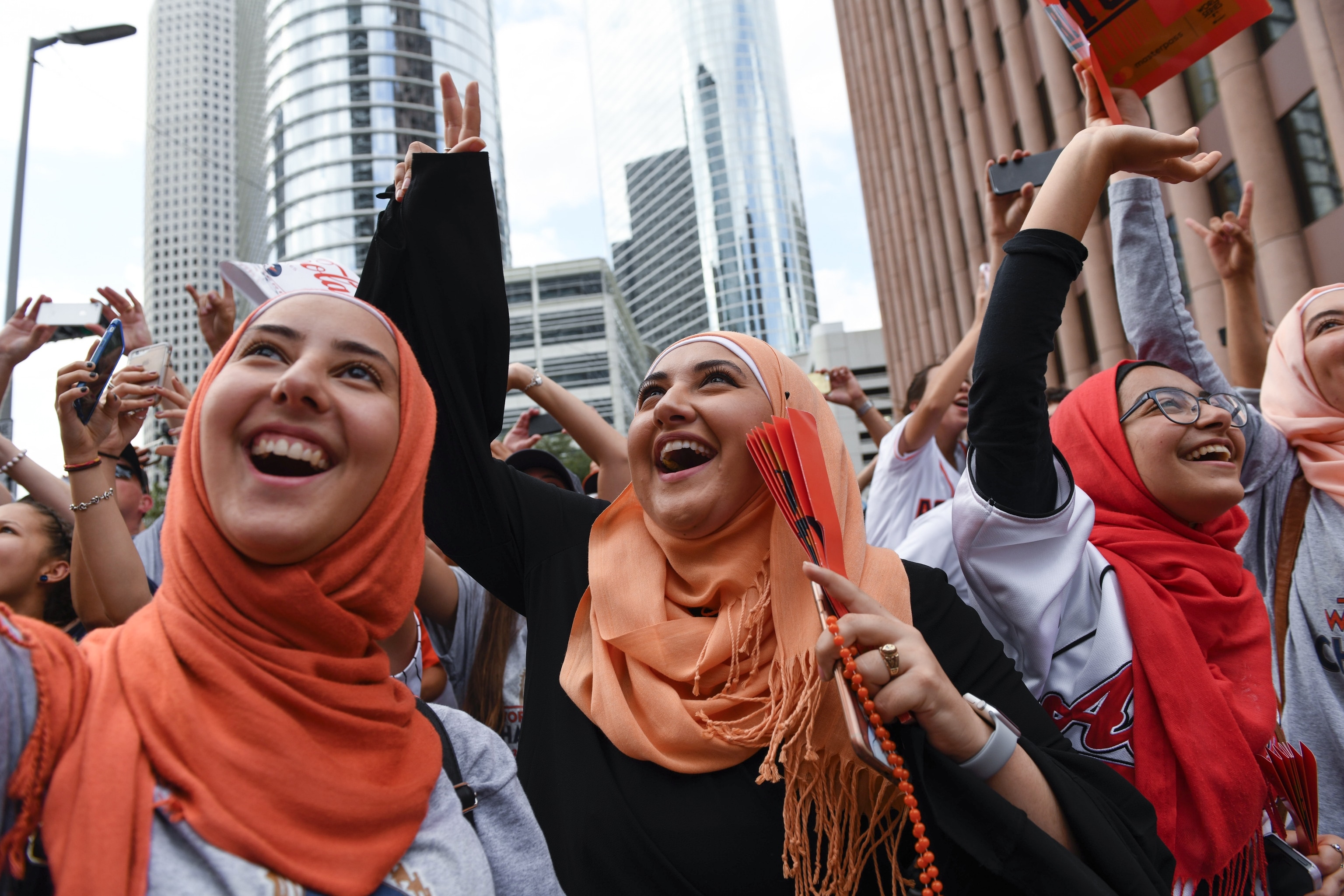 A group of women wearing various shades of orange hijabs, smile brightly and wave.