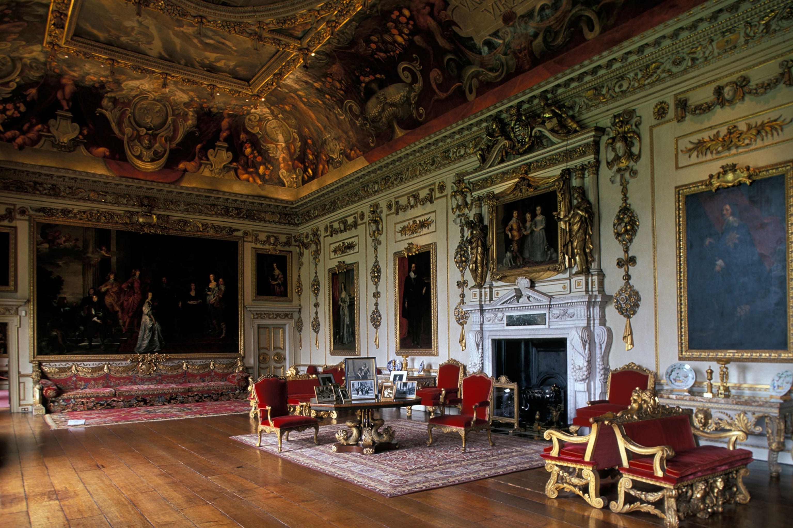 A large, elaborate room with wood floors, high and painted gold adorned ceilings, velvet and gold furniture, paintings on the wall and a large fireplace.
