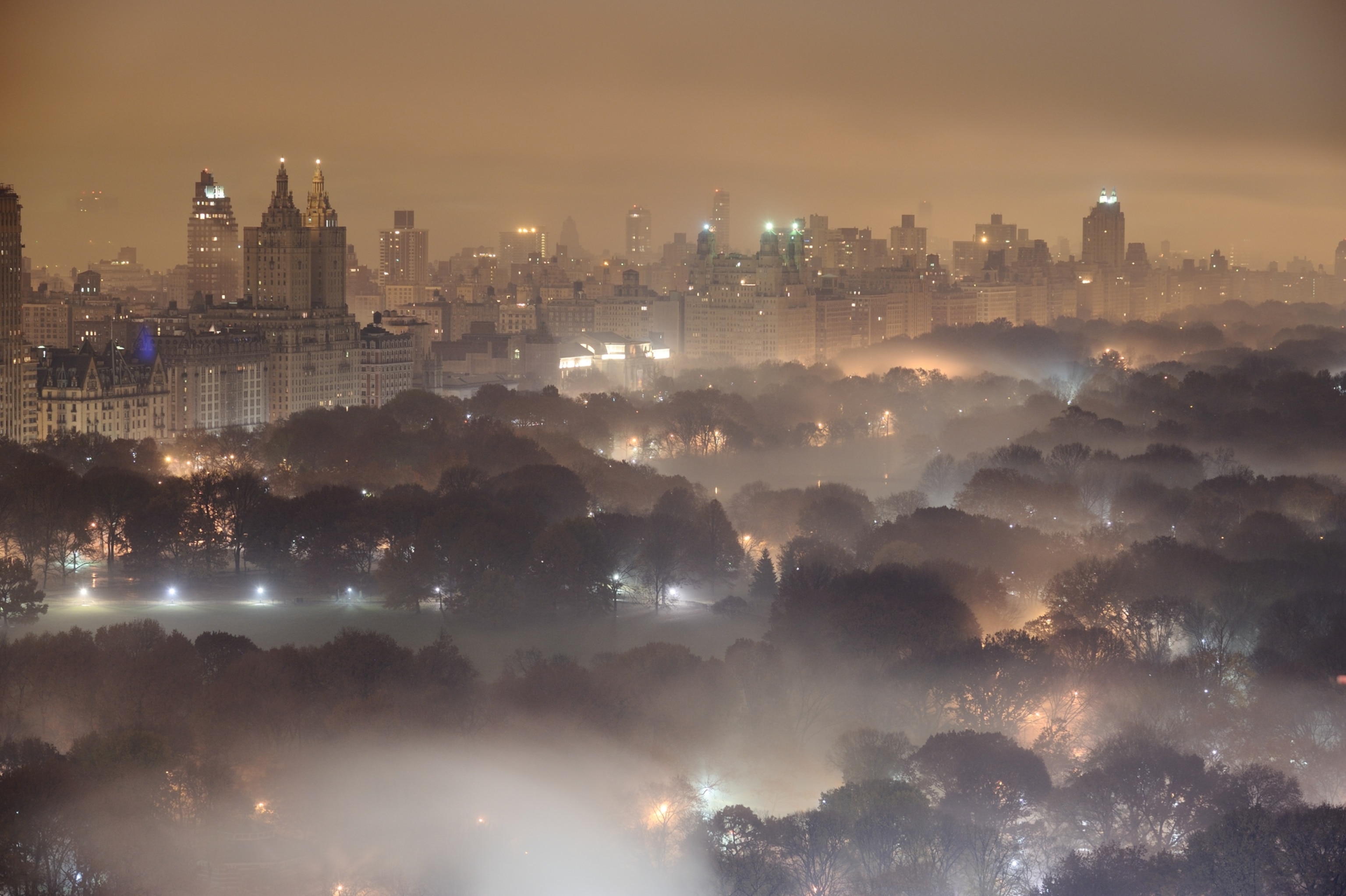 Light pollution and fog combining to blur a New York City skyline at night. Central park sits eerily blanketed by fog in the foreground.