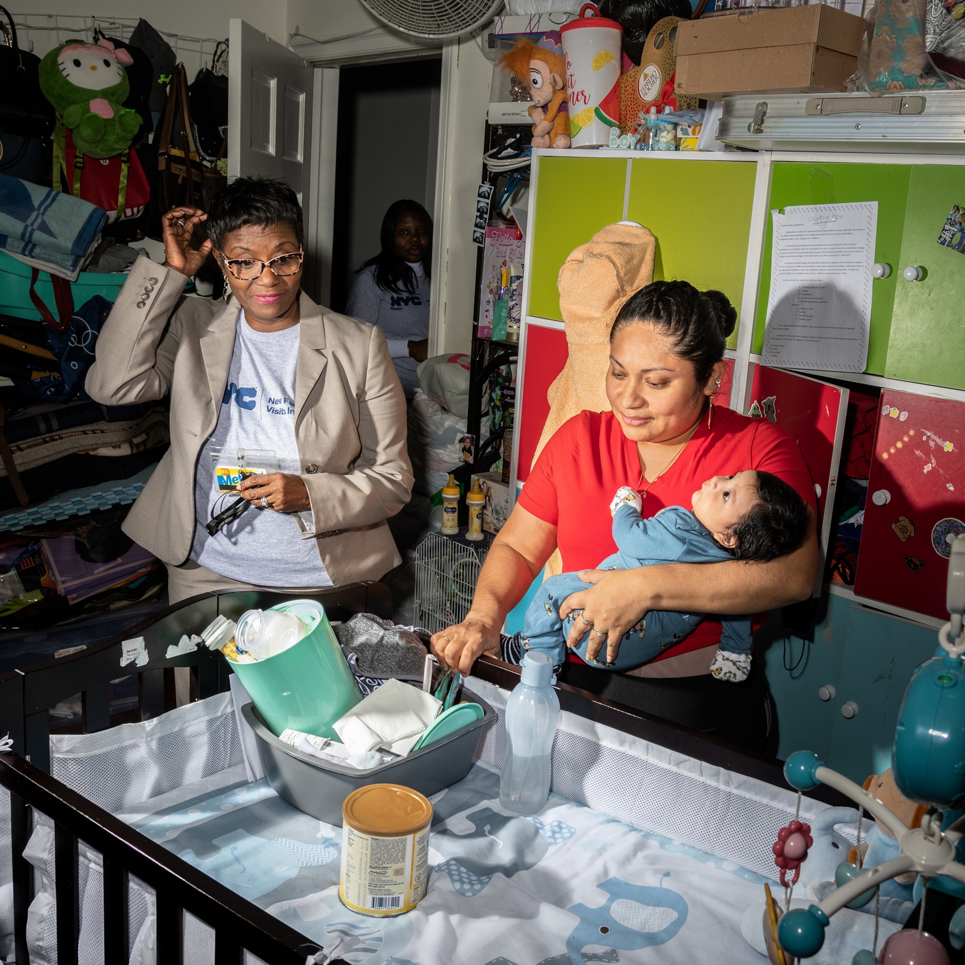 A women wearing glasses and a blazer stands next to a woman in a red top holding her baby to her chest as they stand over a baby's crib.