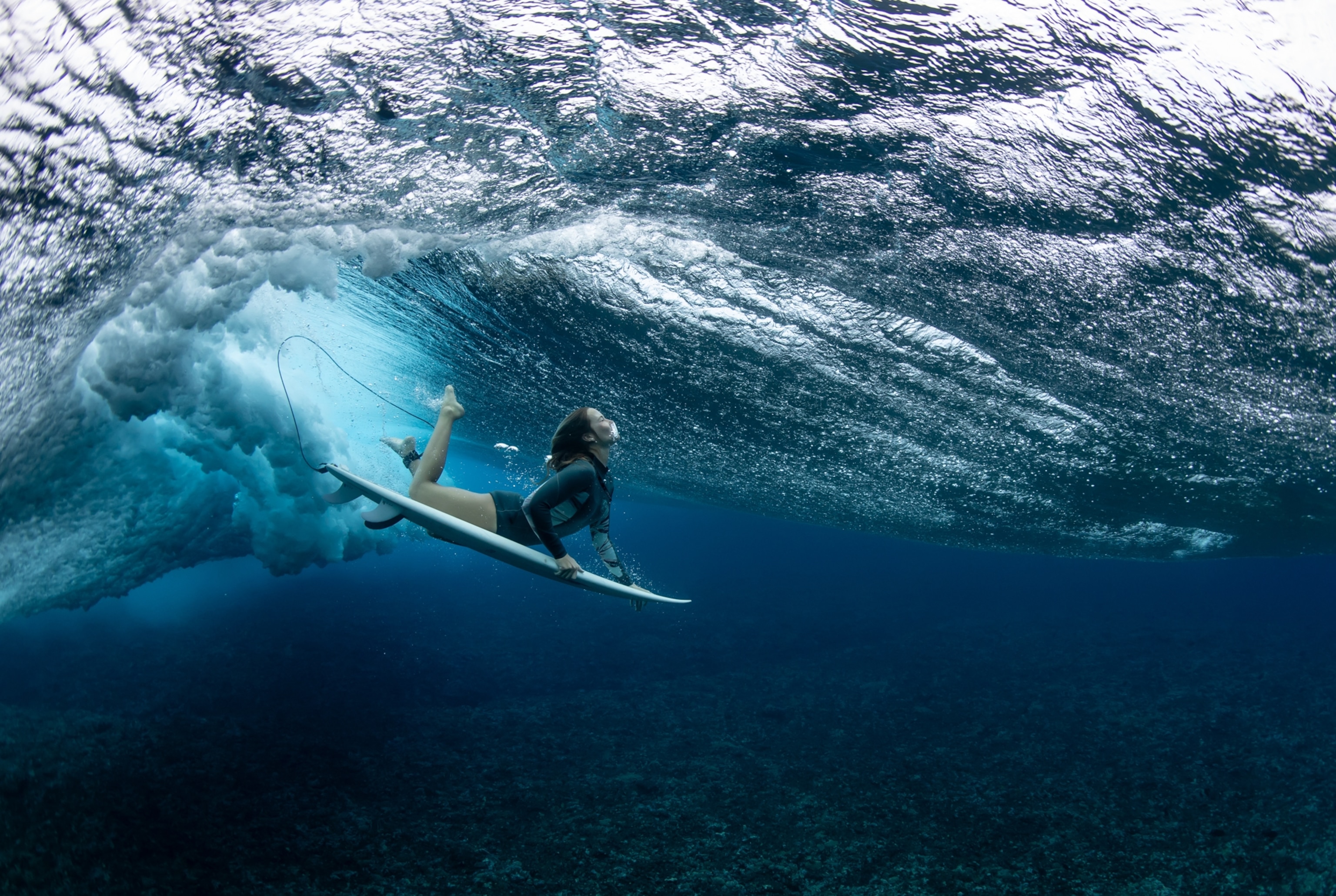 An underwater view of a surfer with her board, diving beneath a wave, which rolls like a blanket of water above her.