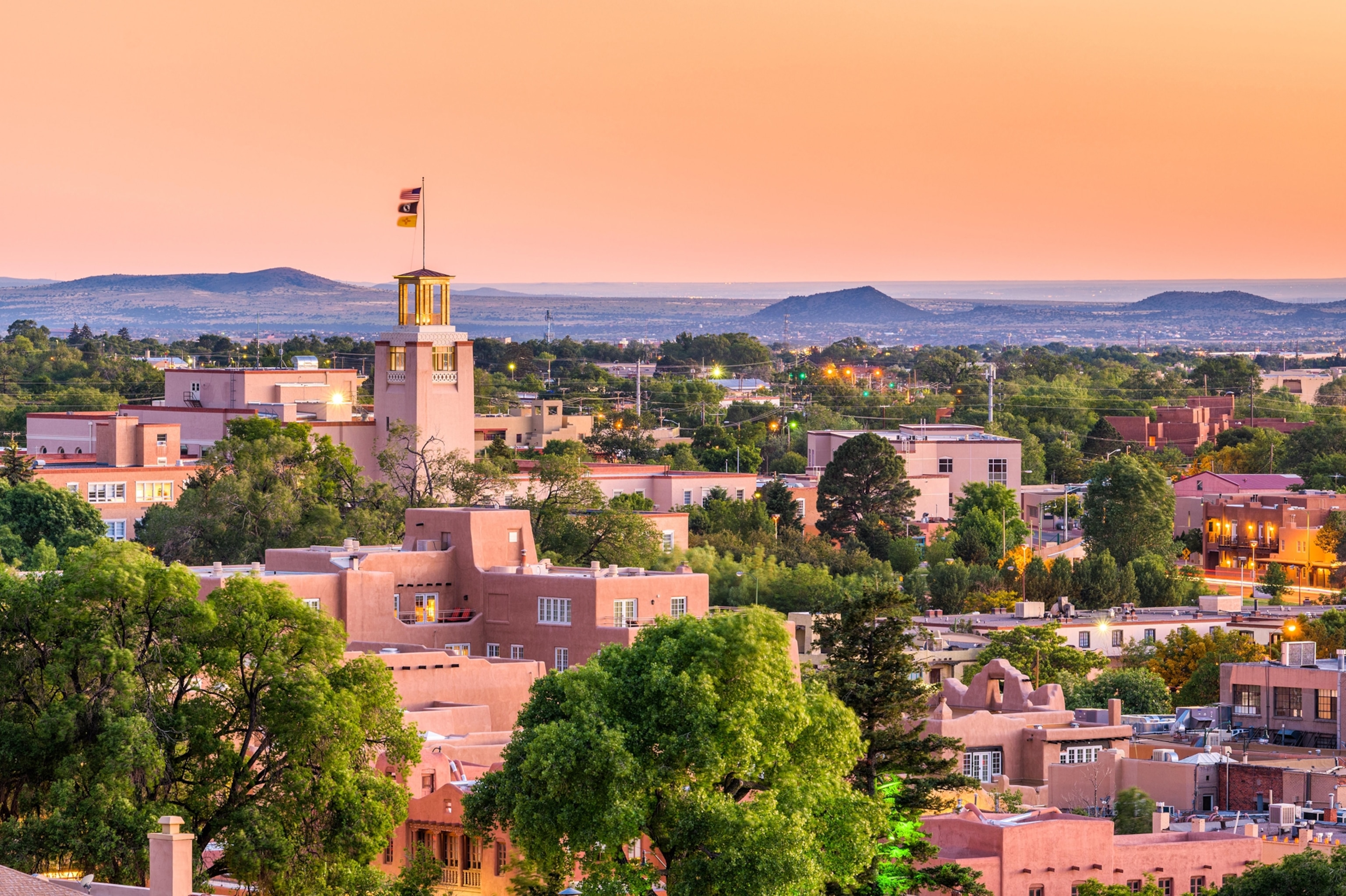 Light pink buildings of Santa Fe, New Mexico are seen amidst scattered trees. It appears to be approaching evening as the streetlights are on. The sky is a light orange. There are low rise mountains in the background.