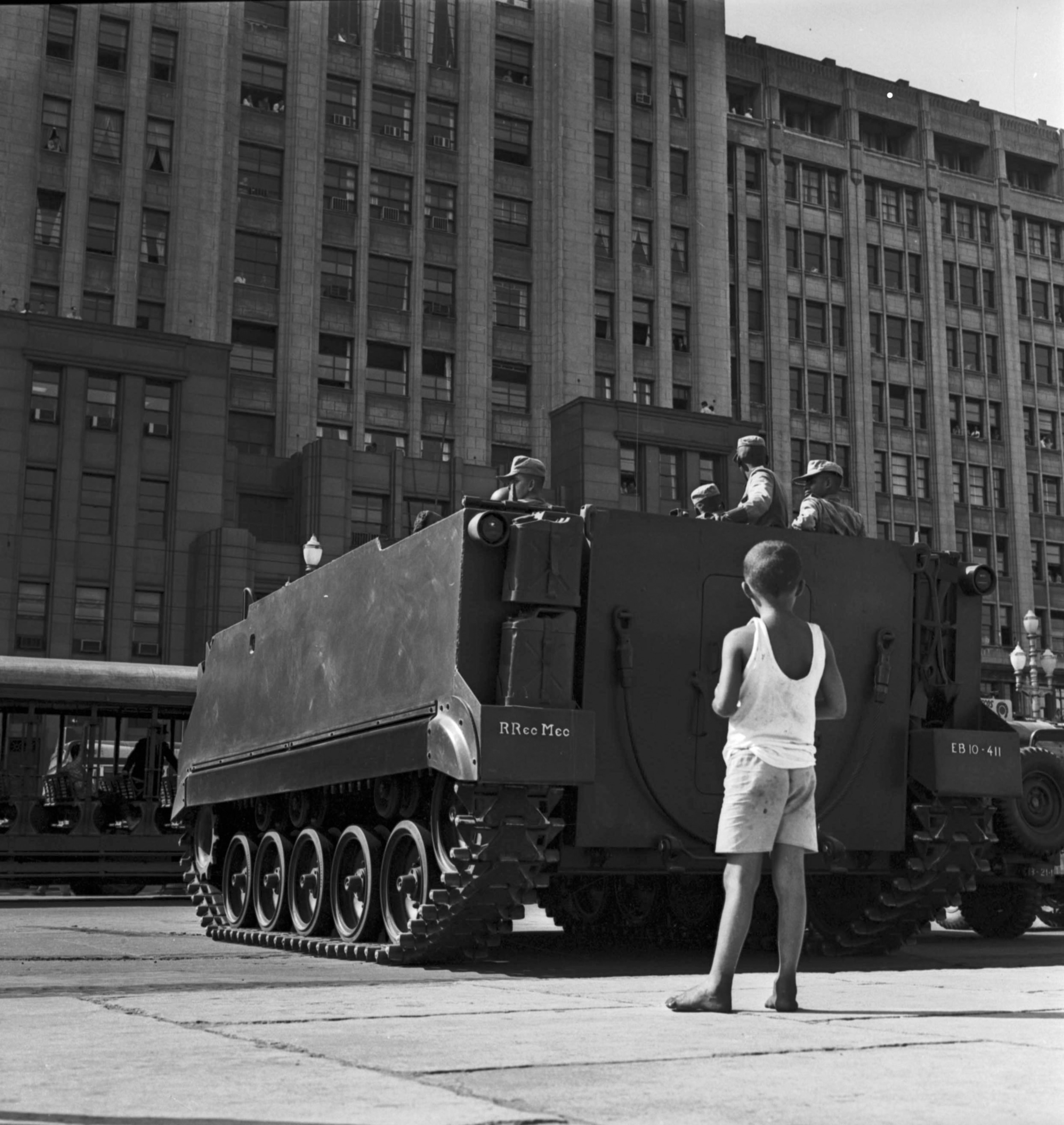 A Black and white photograph shows a young boy standing as a tank is driven on the street.