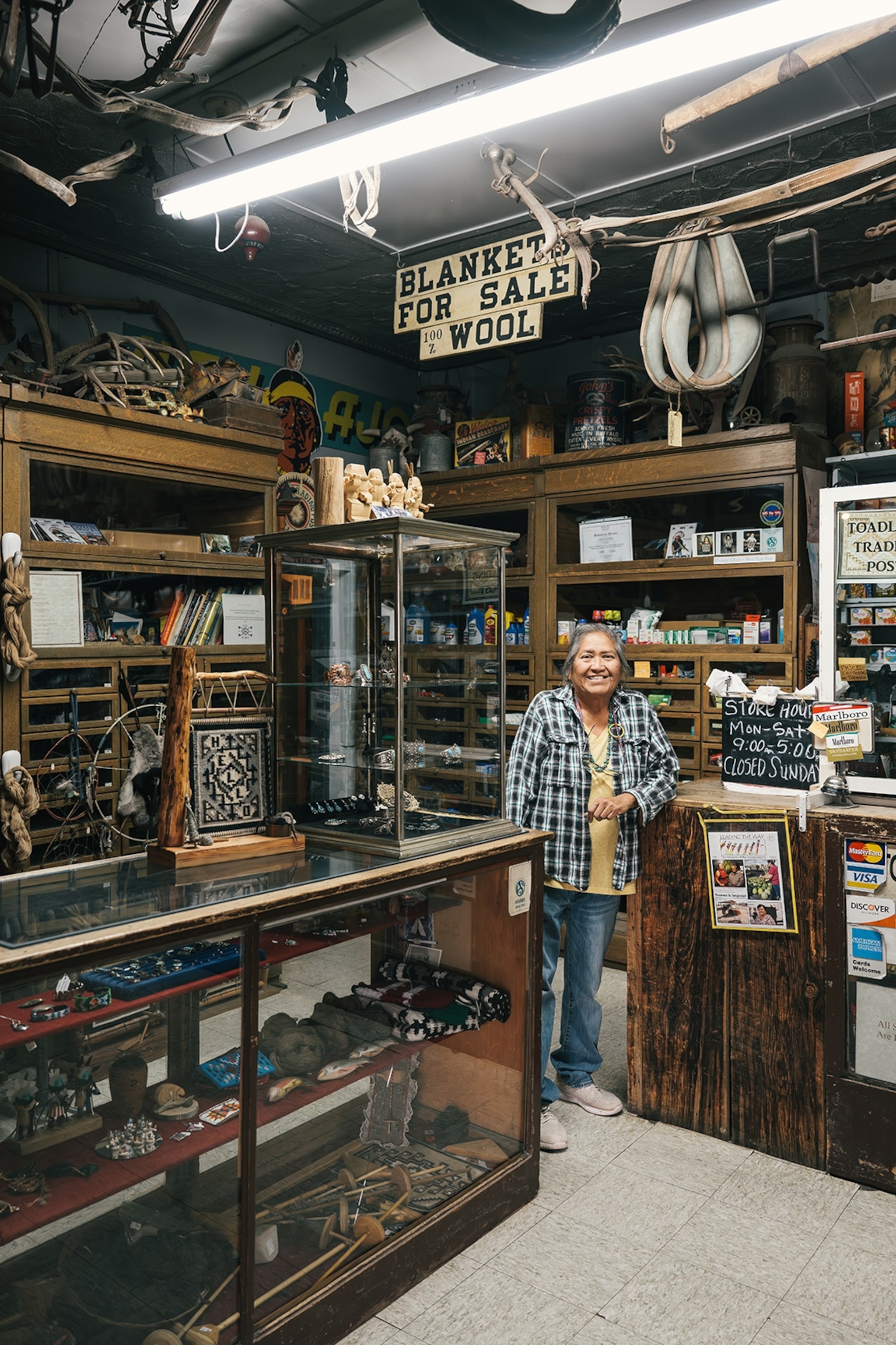 An elderly, smiling woman standing in her LED-lit, wooden trading shop.