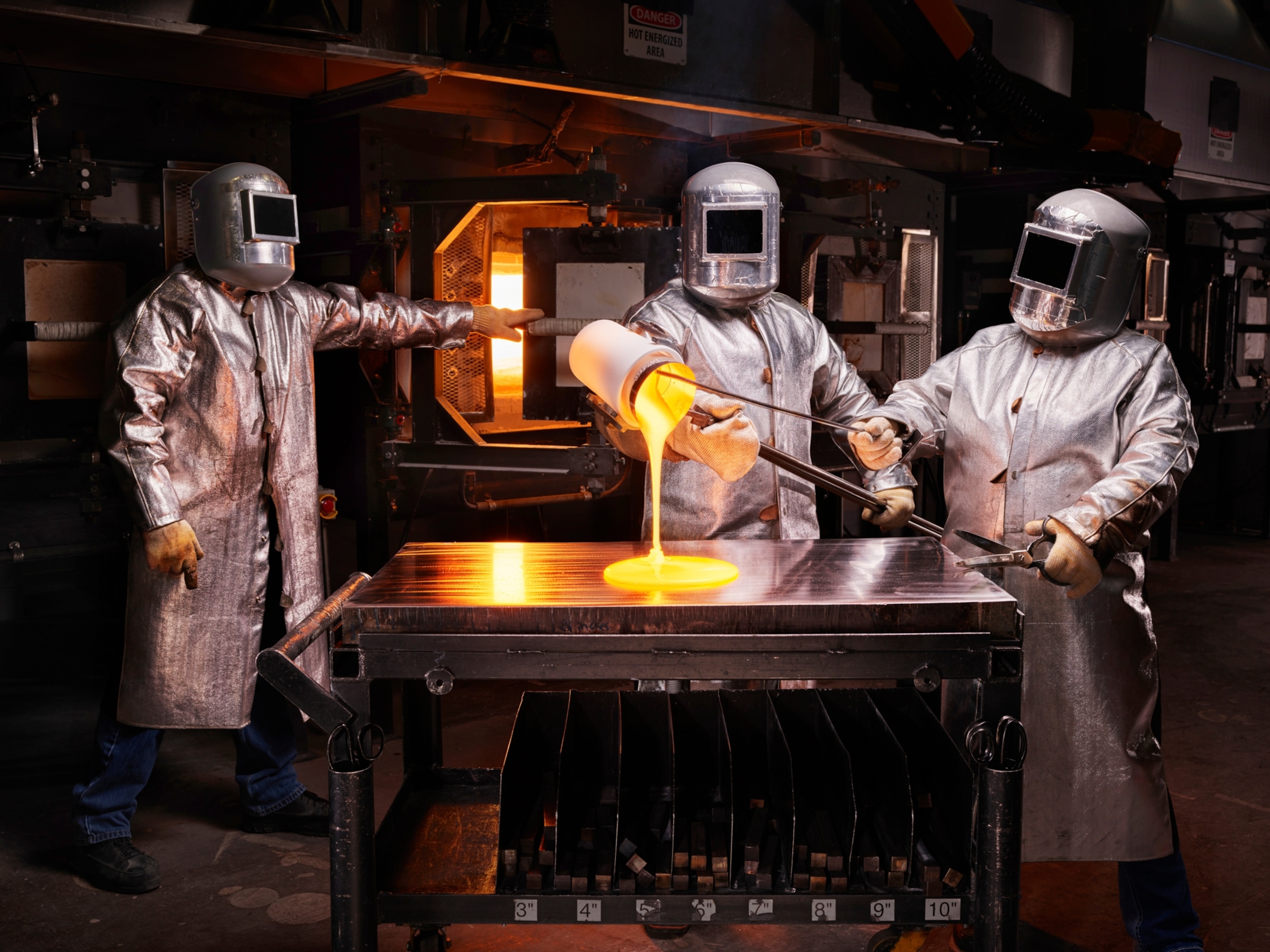 three individuals in silver suits stand around the table with a glowing orange furnace behind them. One technician pours out bright orange liquid glass onto the flat silver table.
