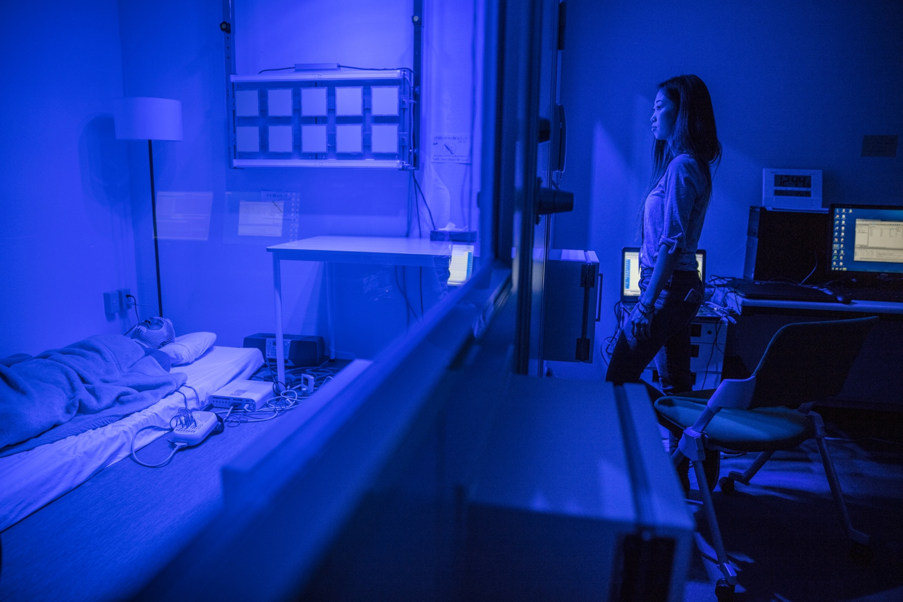 a woman looking at a man in a sleep study behind a glass wall, they are cast in blue