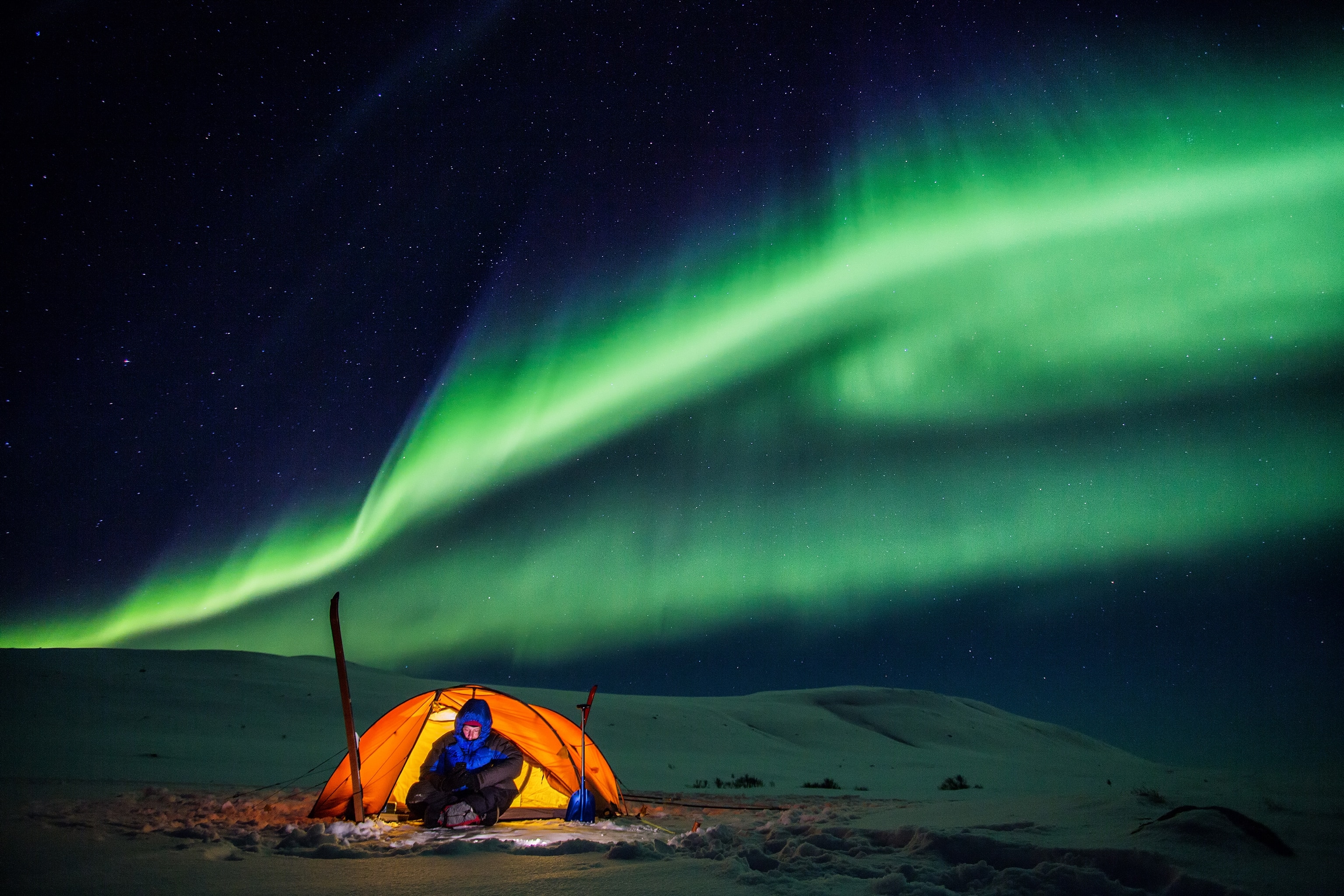 A man in an orange tent and blue coat, under the northern lights.