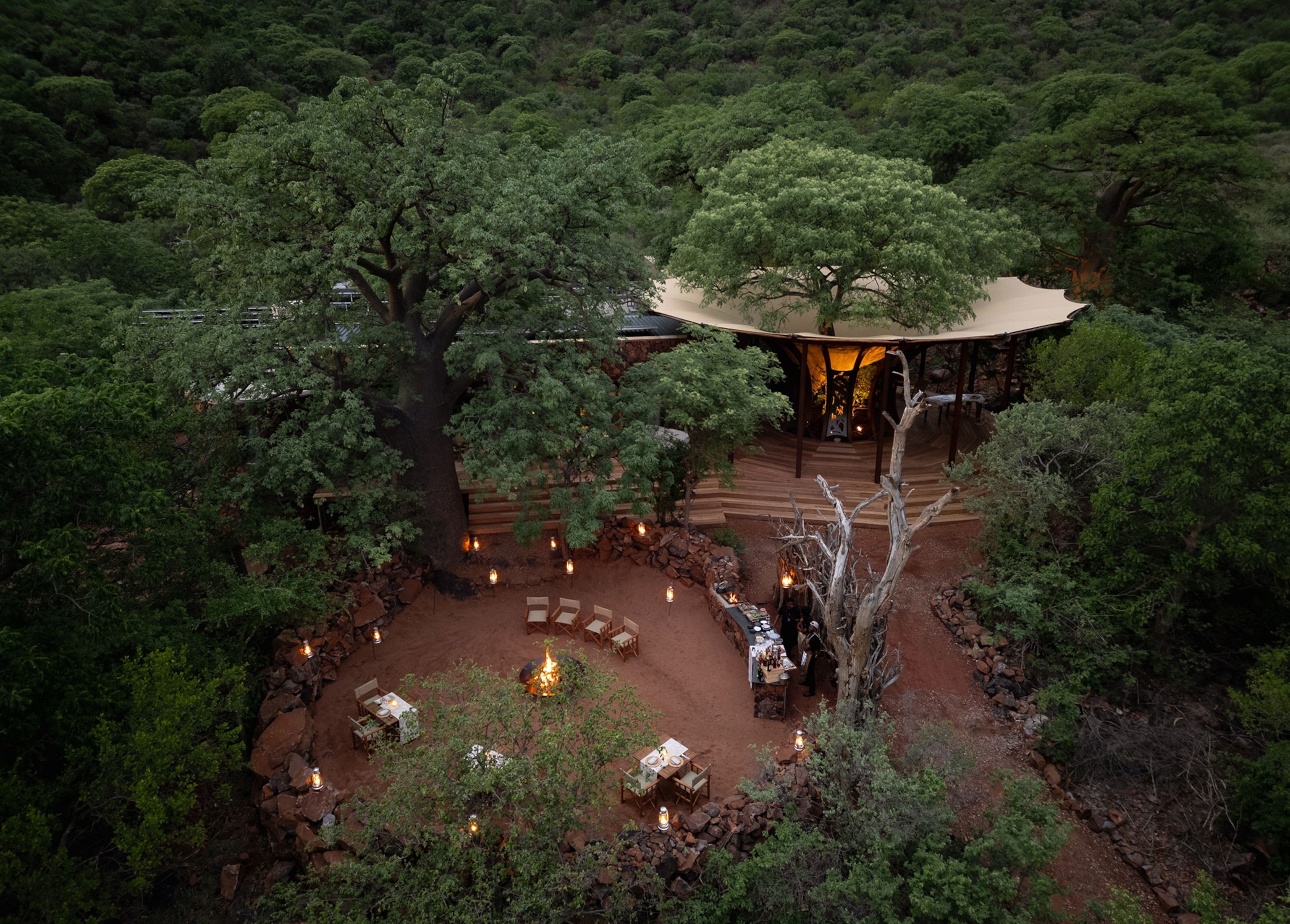 Aerial view of lodge surrounded by trees with a campfire sitting in open space next to building structure.