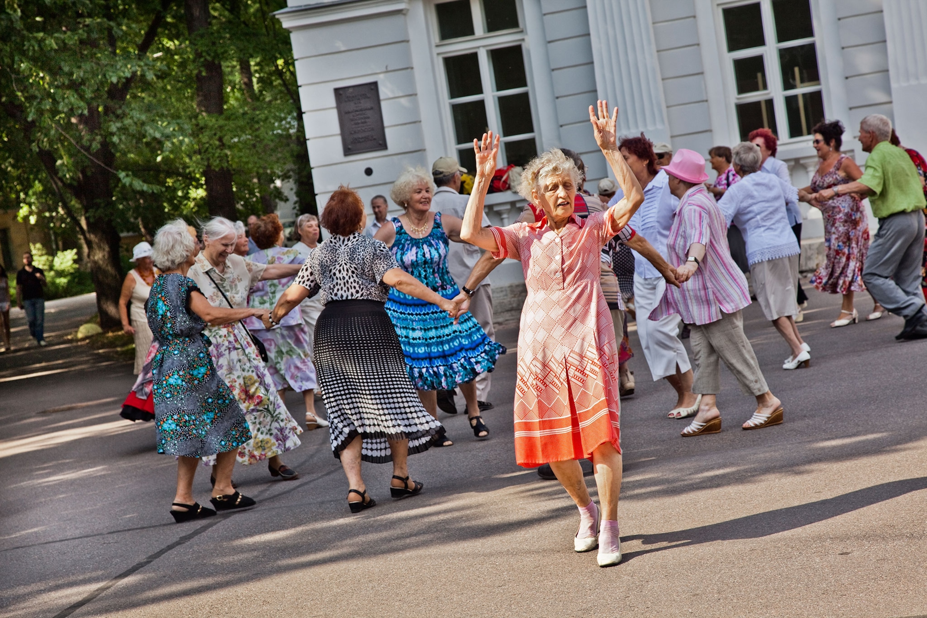 An elderly Russian woman, wearing a bright pink and orange patterned dress, joyfully dances on a sunny summer afternoon in a park with a group of women dancing in the background.
