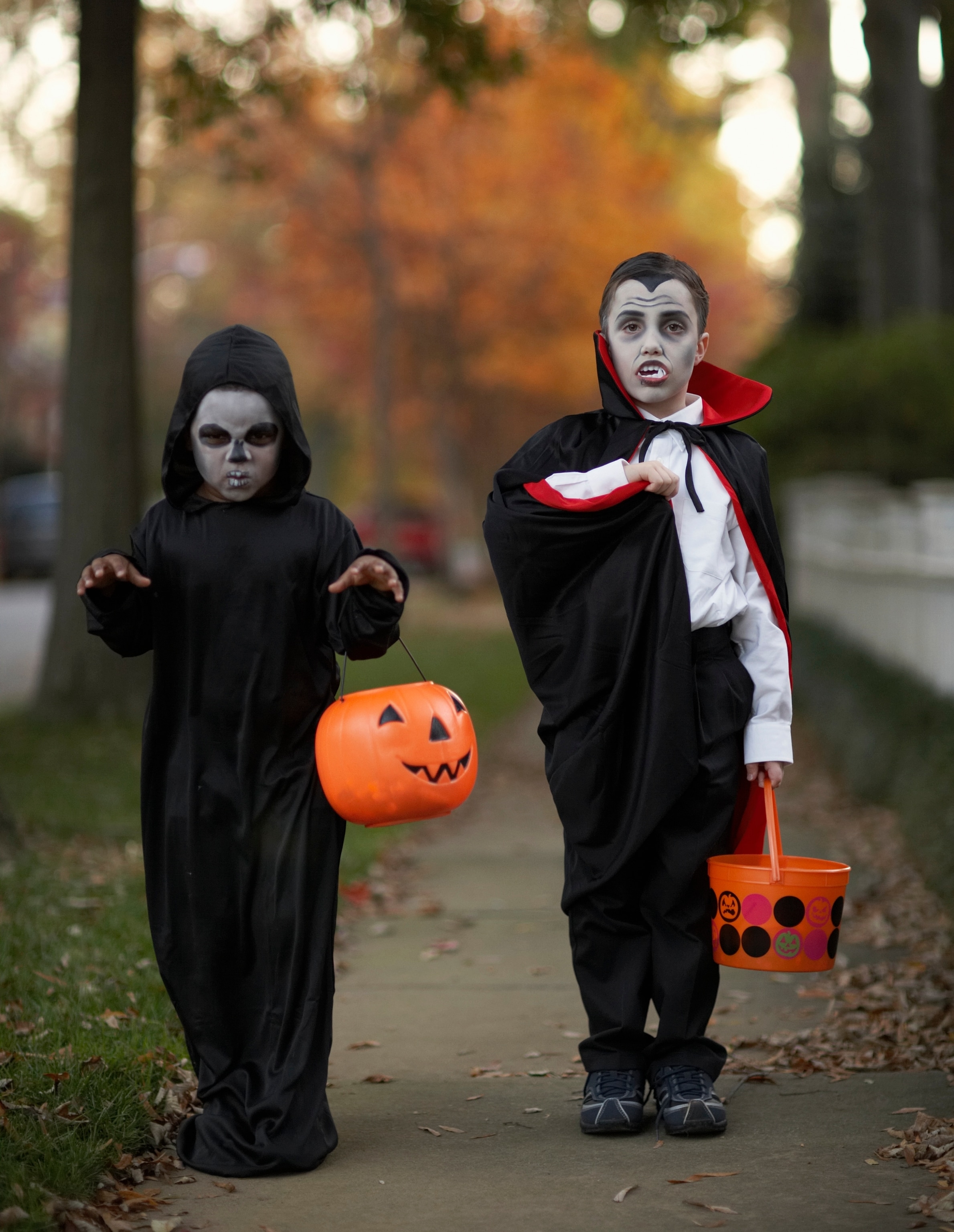 Two grade-school-aged boys wearing Halloween costumes and face paint stand on the sidewalk in a neighborhood while trick-or-treating.