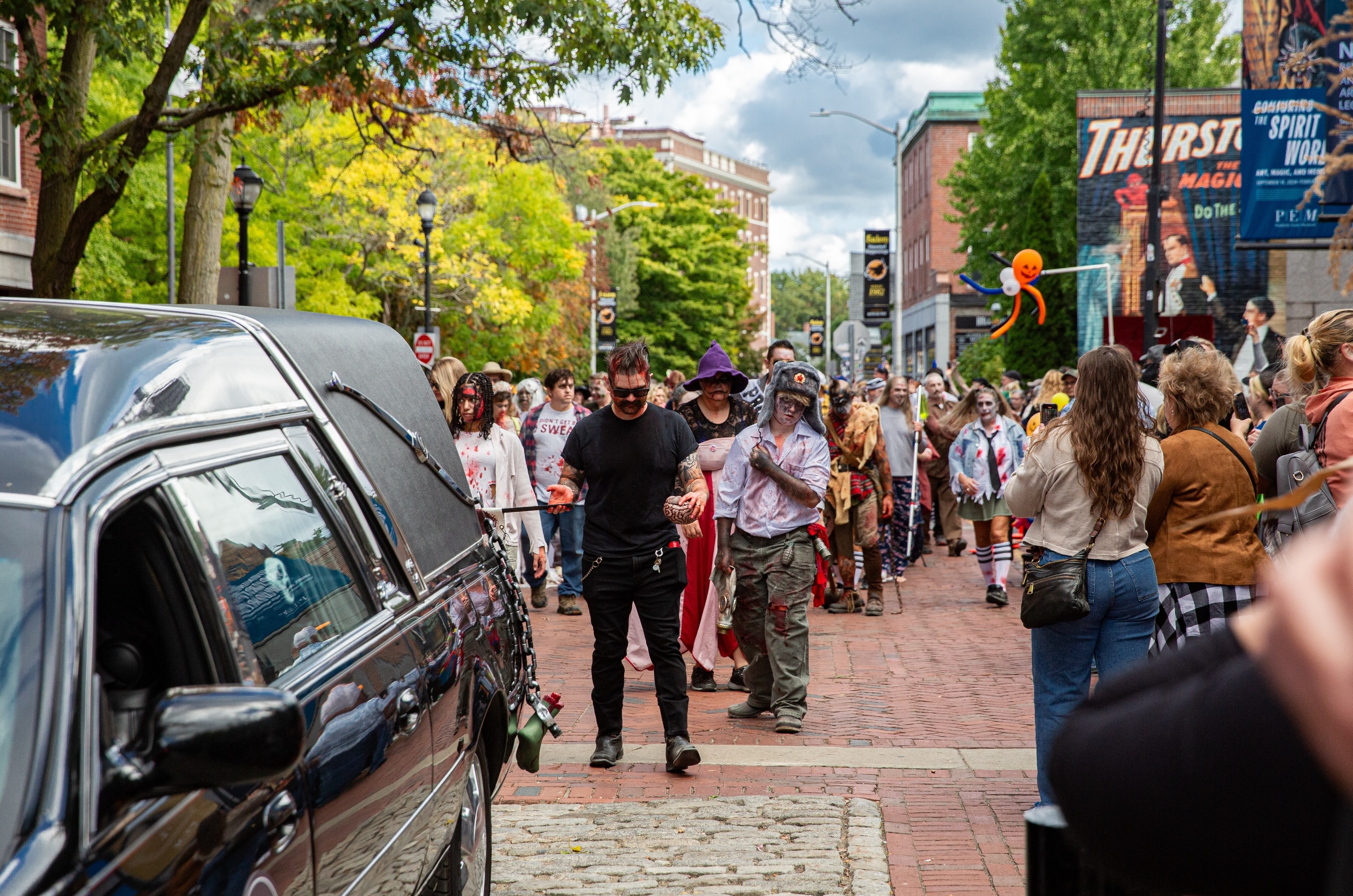 A group of people dressed in zombie costumes walks down a street as part of a Halloween parade.