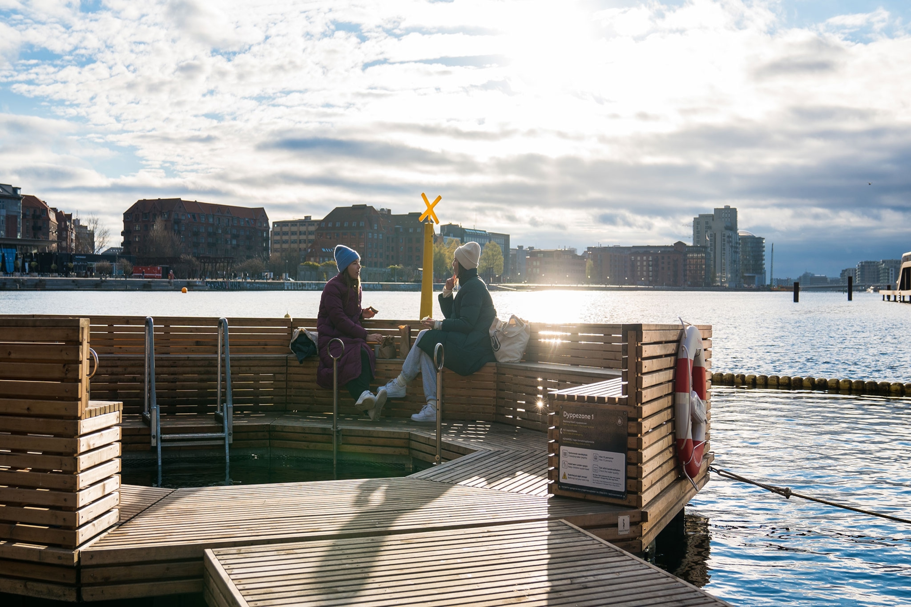Two women sit in warm coats on the edge of the harbour in Copenhagen.