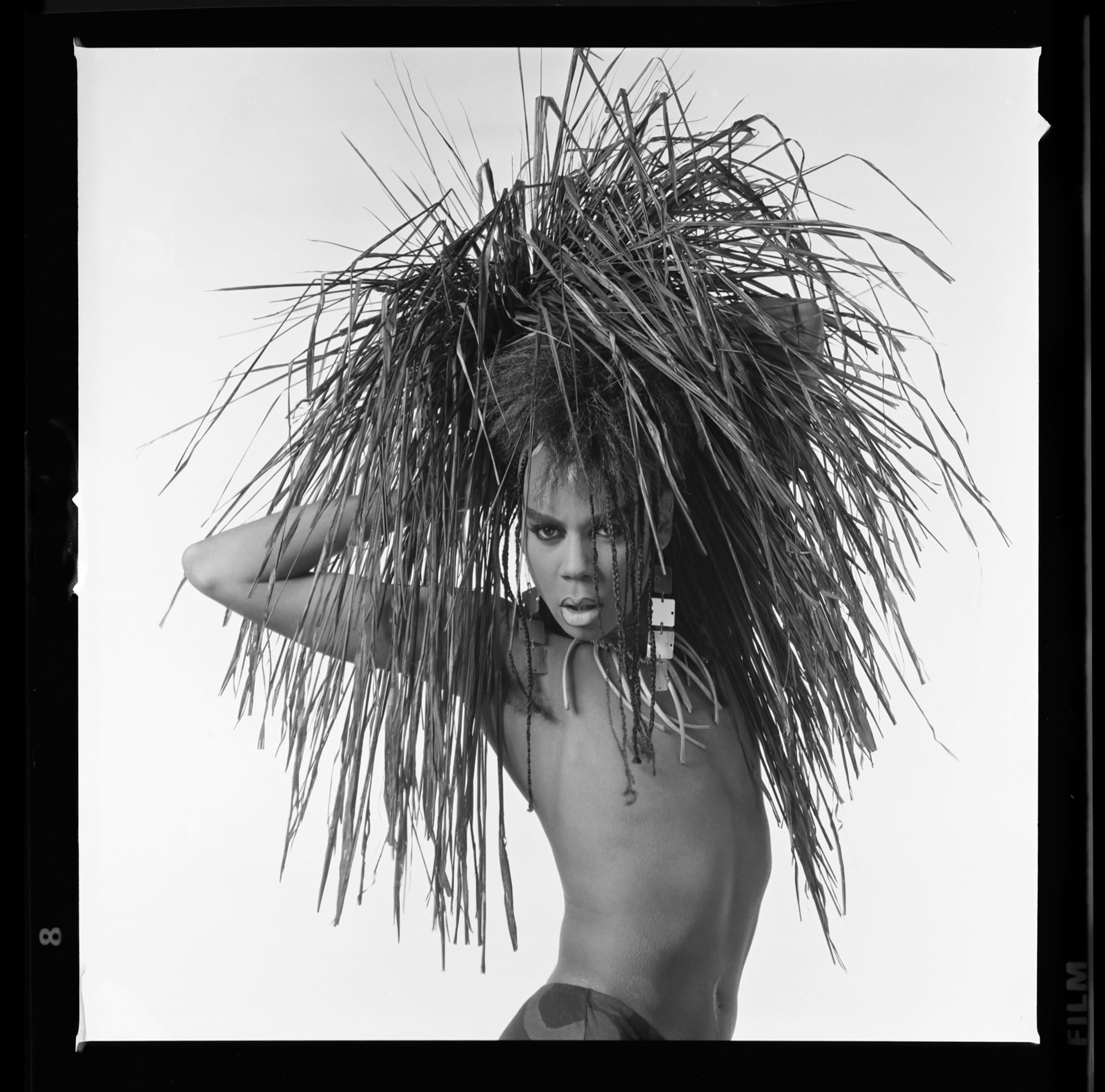 A black and white image with a strong pose and gaze directly into the camera holding reeds.