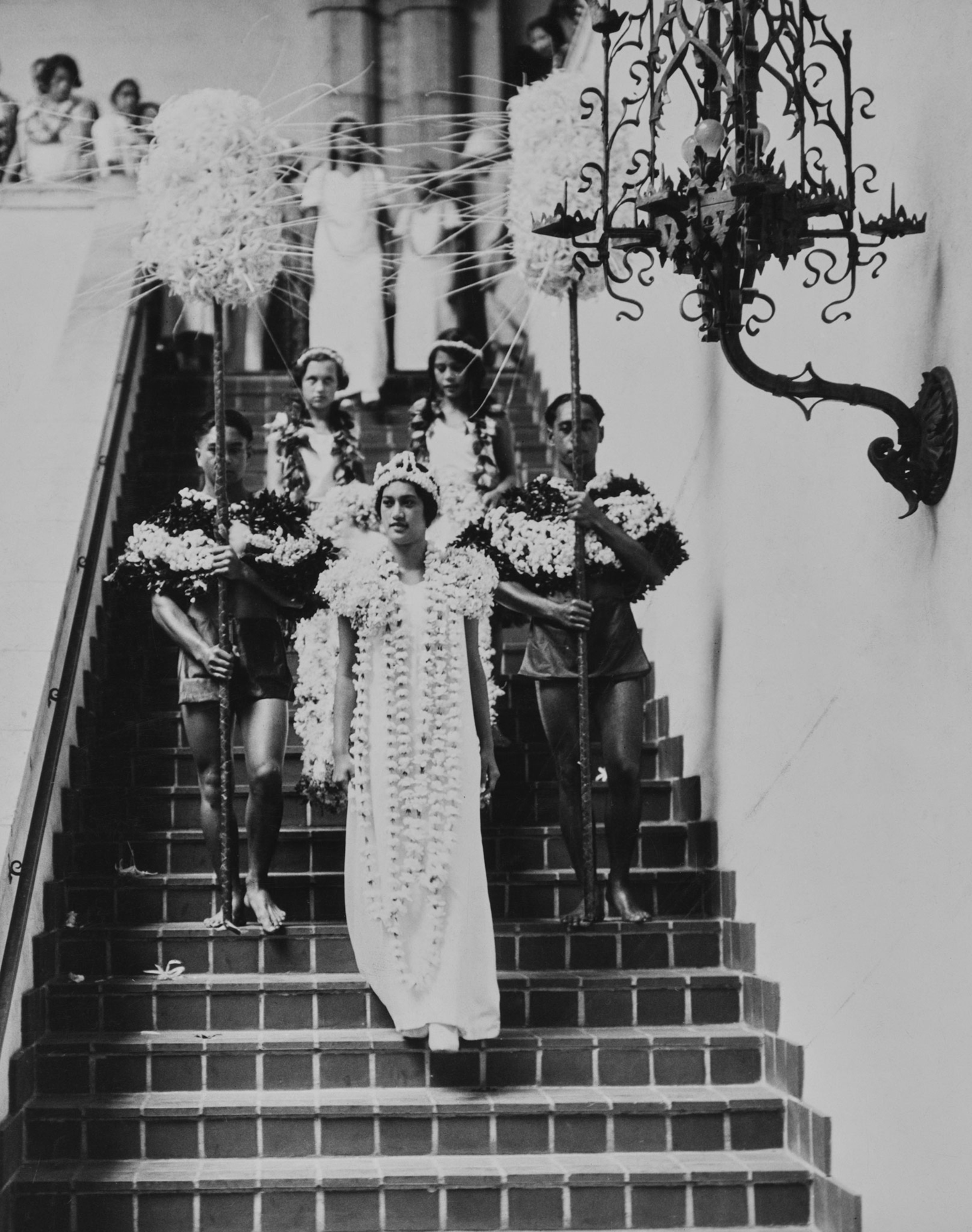 A woman dressed in white adorned with flower leis and a crown walks down stairs, followed by 2 men and 2 girls also wearing flower leis