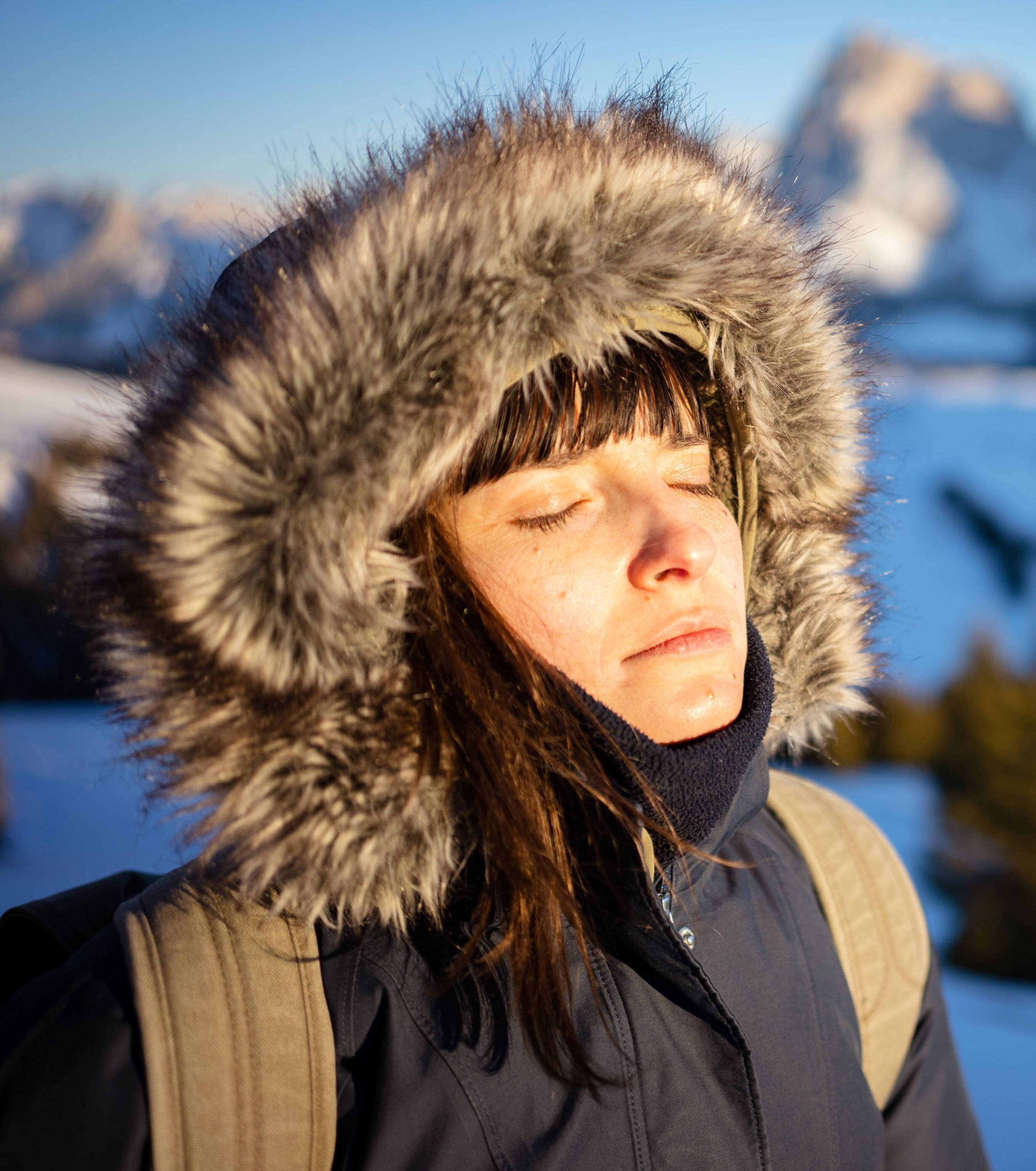 a woman with a black winter jacket and a large, fur trimmed hood exposes her face to the sun with her eyes closed. There are snow covered mountains and trees in the background.