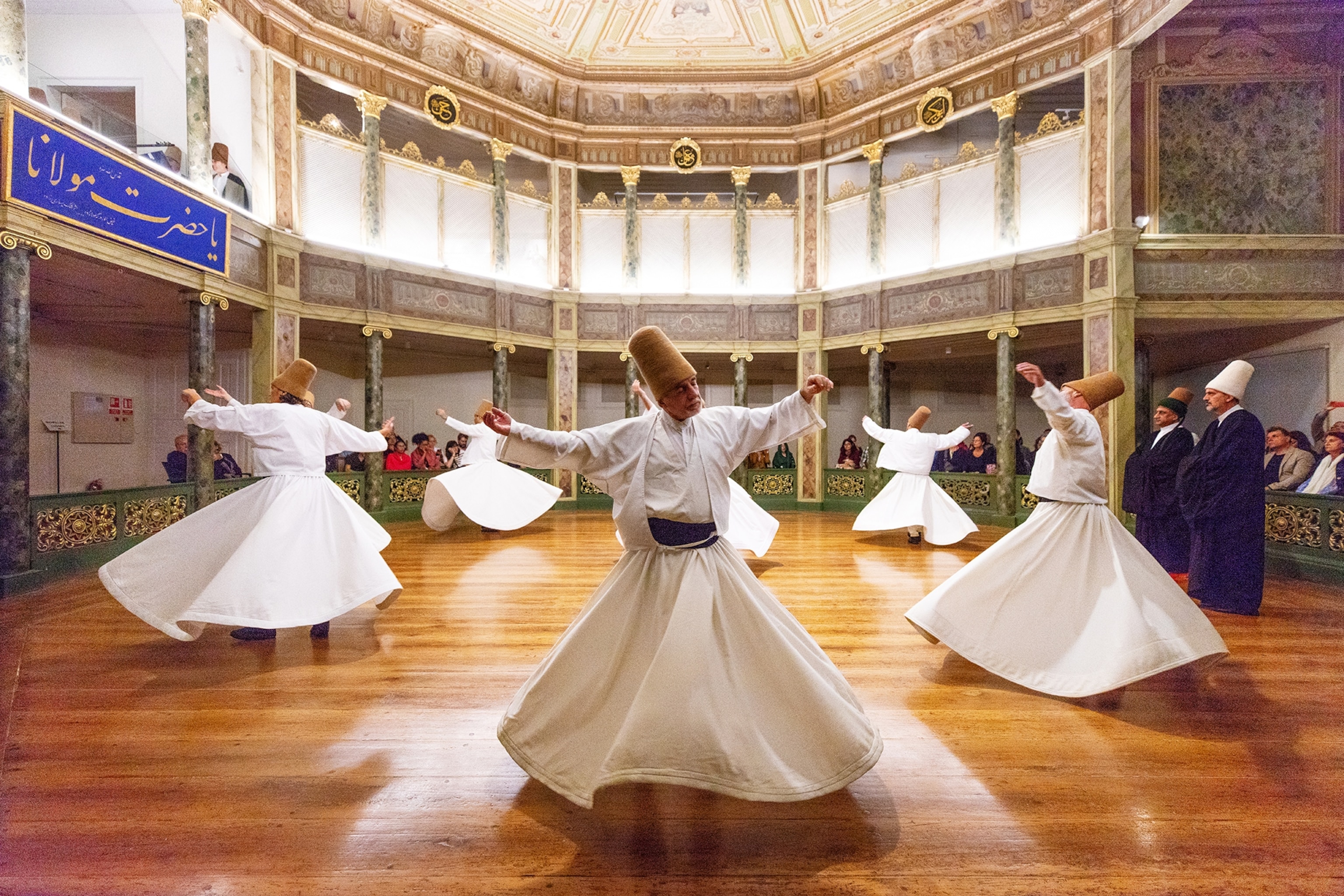 Men wearing white robes dance around a hall with wooden floors and balconies