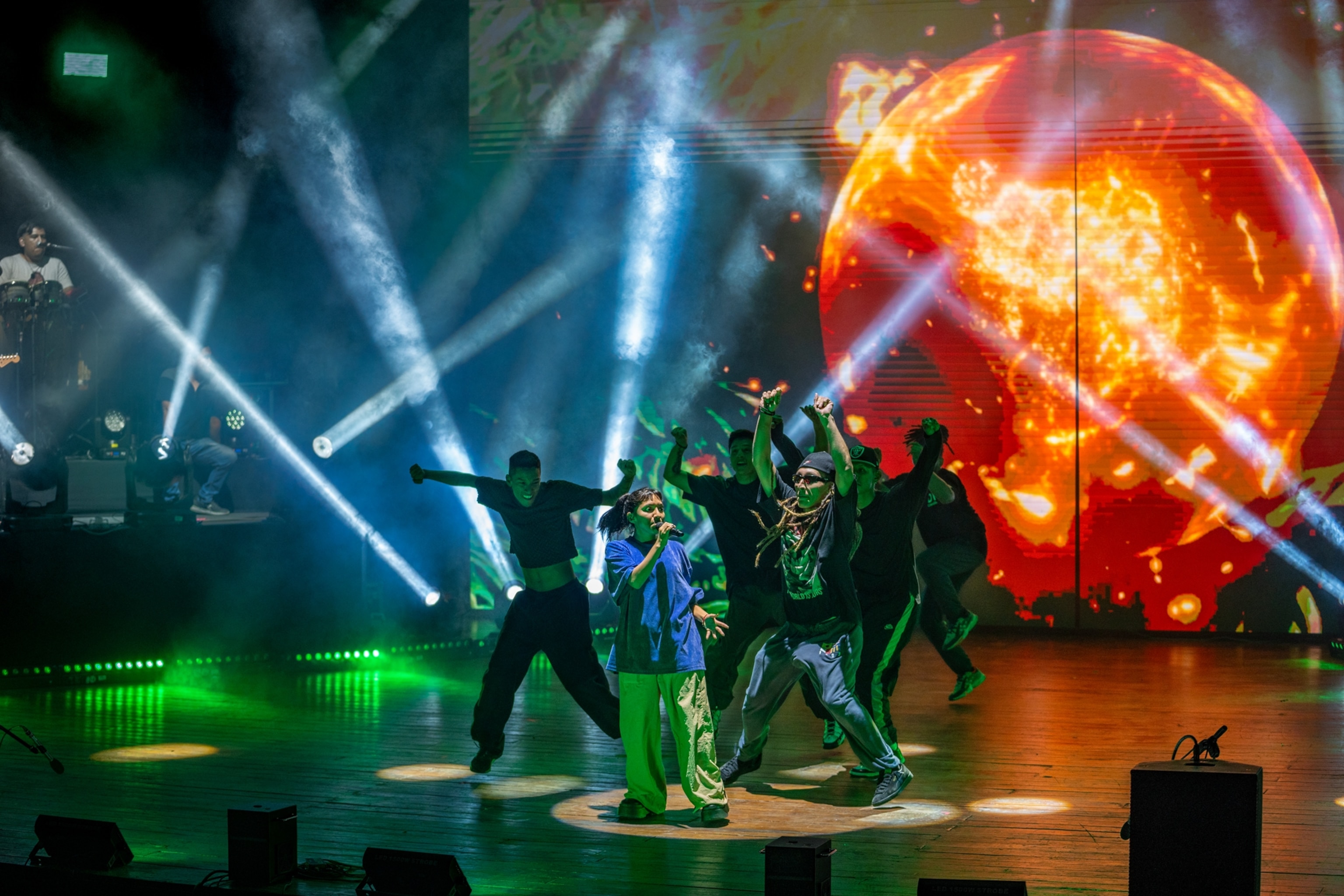 Renata Flores rehearses on stage with several dancers behind her. There is a fiery red back drop and green and blue lights on stage.