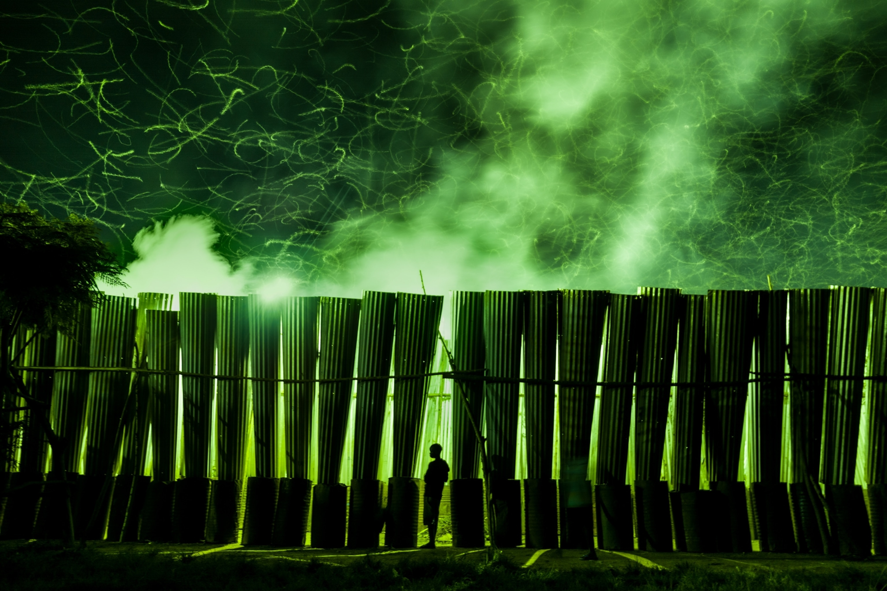 Picture of a man silhouetted in the green light of a line of tall metal nsenene traps as thousands of crickets swirl above.