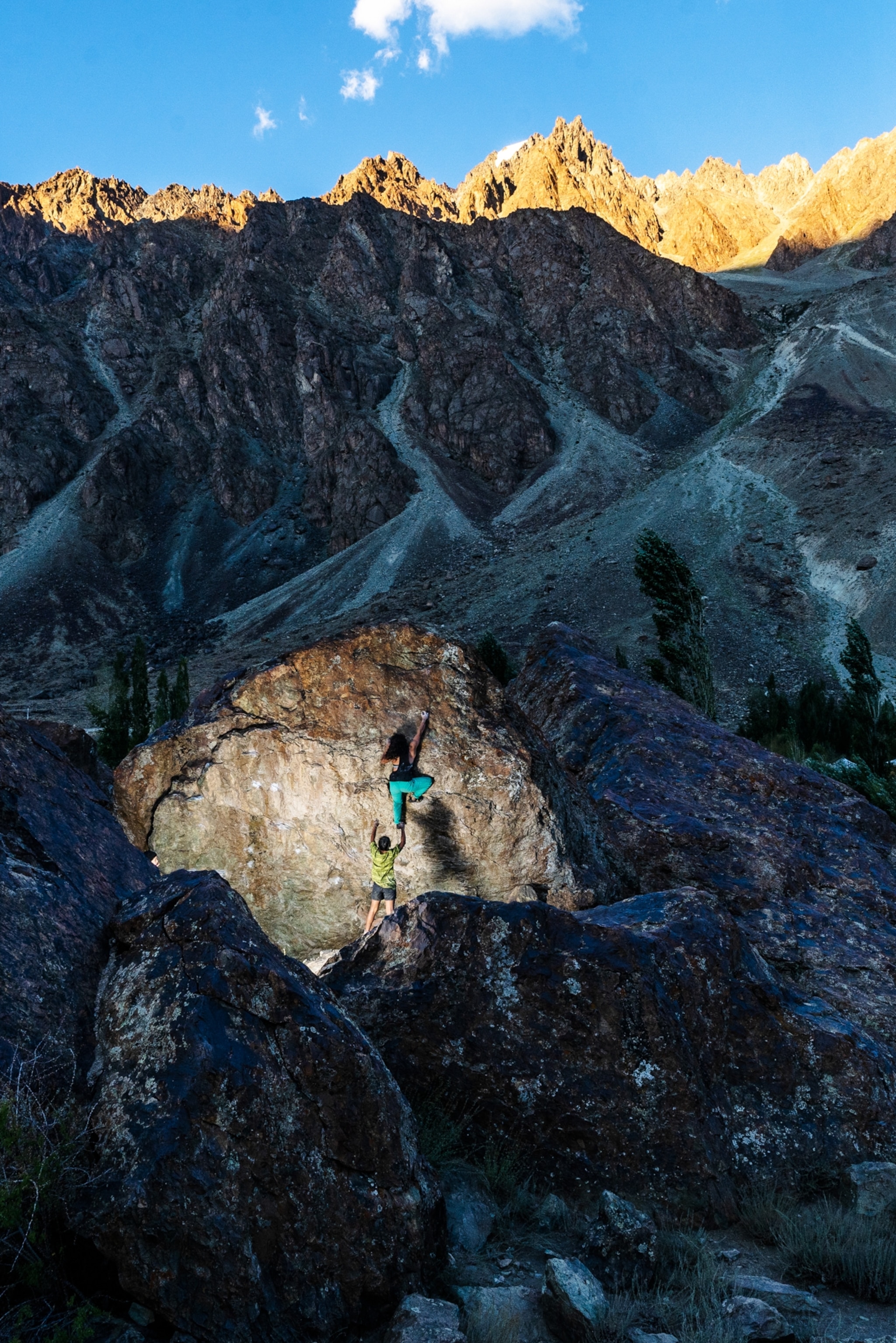A rocky valley with mountain peaks kissed by sunlight in the background. Near the foreground, a camera flash shines on a woman climbing the sheer face of a boulder as another woman stands by as a spotter.