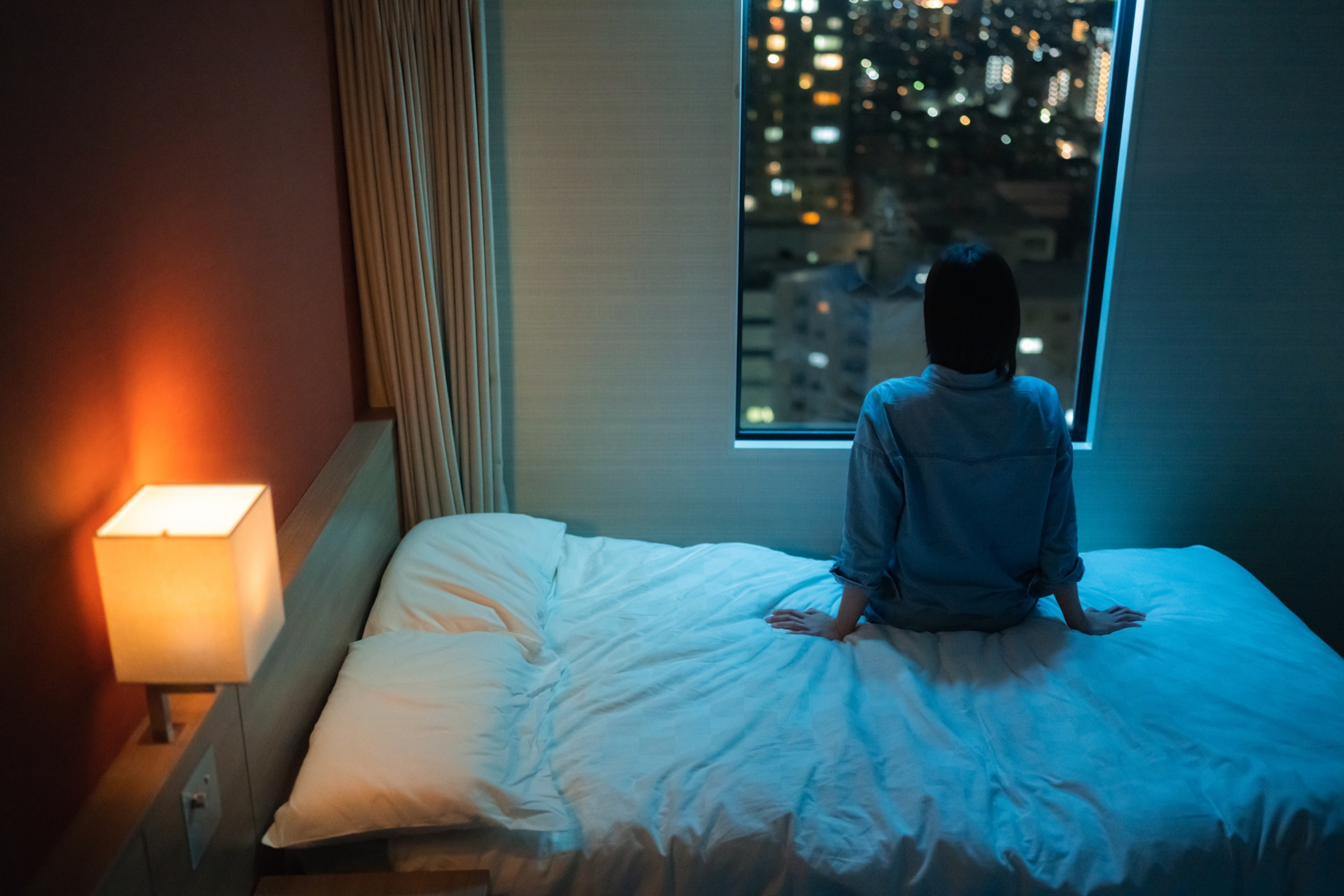 A woman sitting in bed, seen from behind as she looks out the window at a city at night.