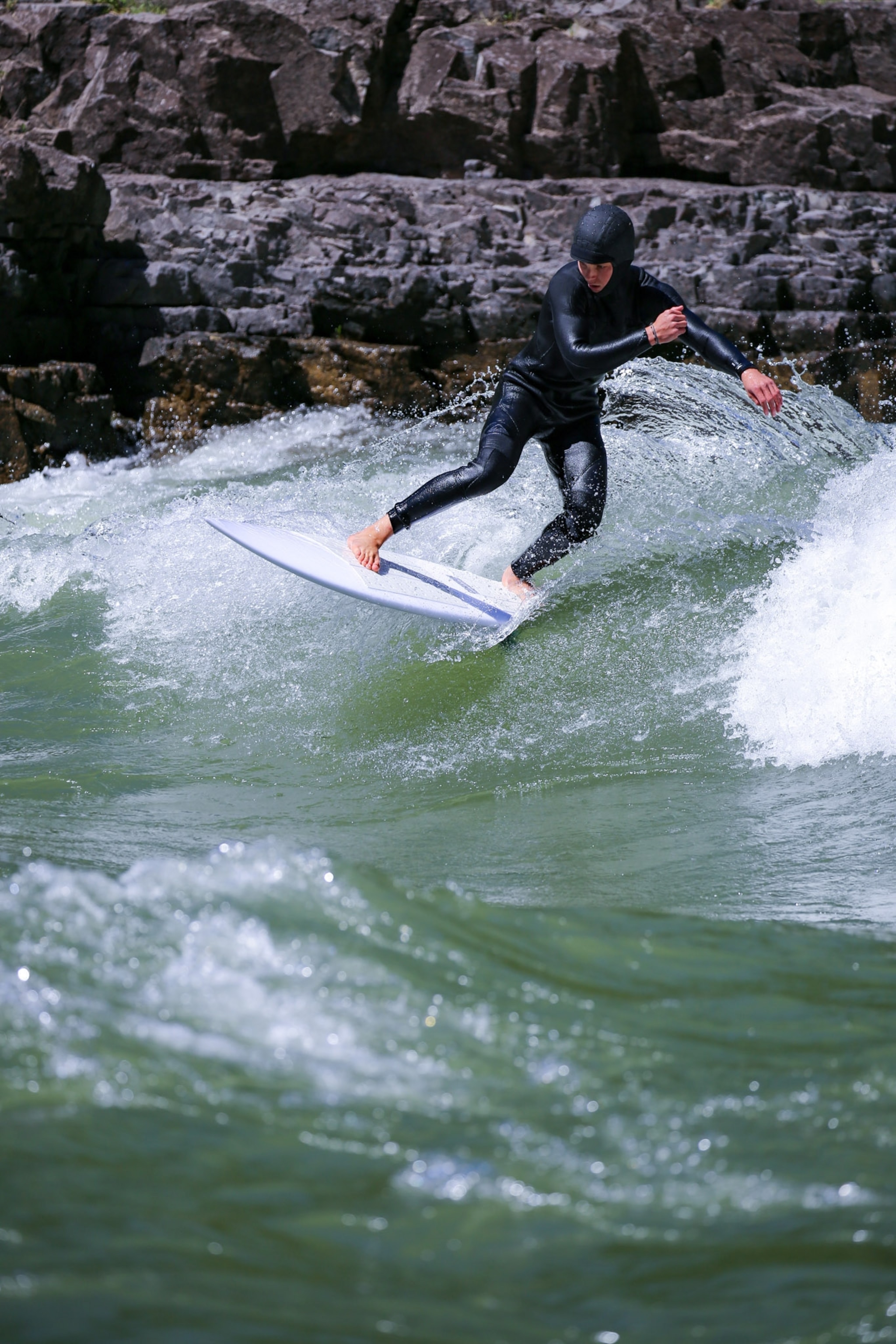 A person in a black suit surfing in a river rushing waters and dark rocks behind her in the frame.
