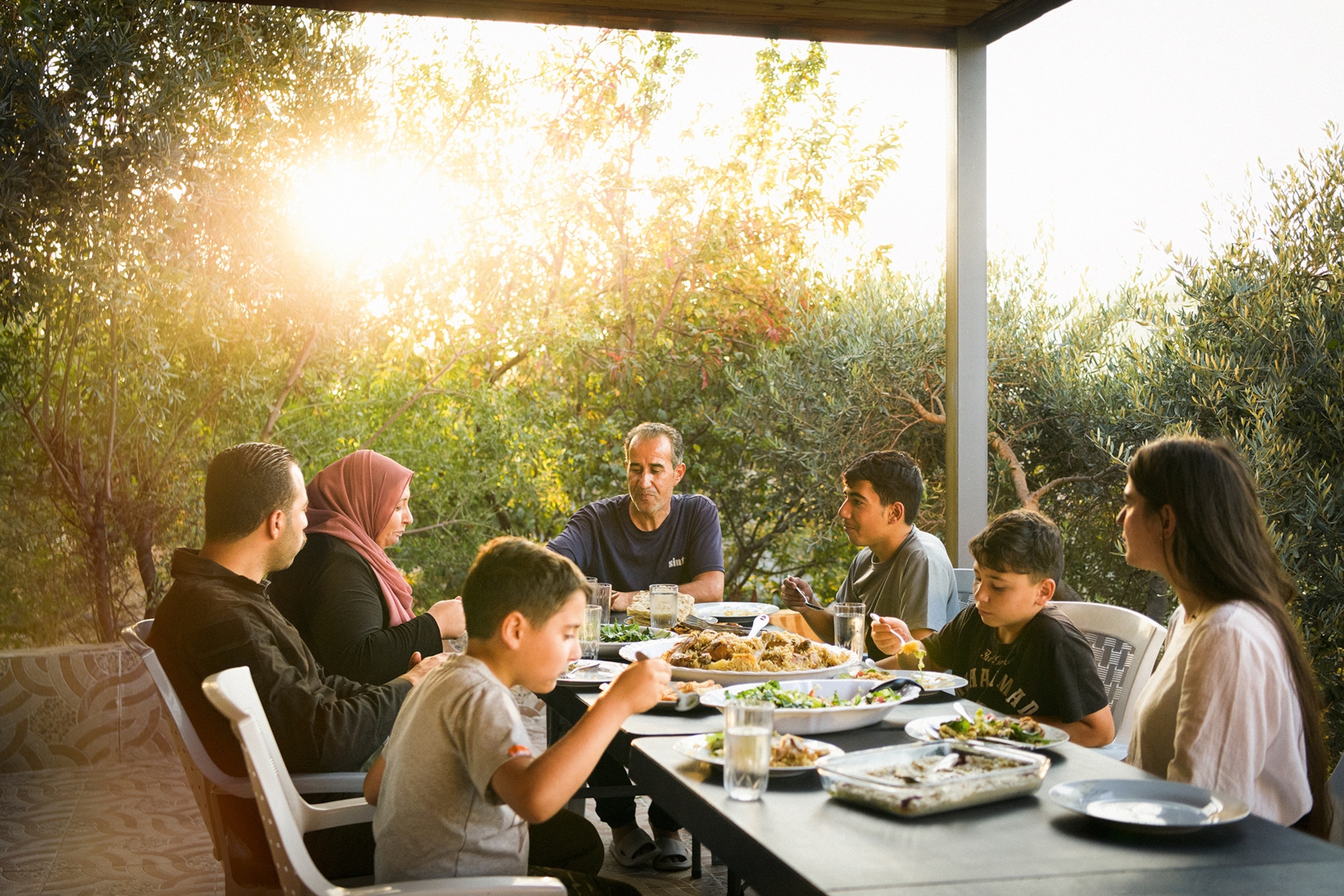 A family enjoying dinner on a long table as the sun is blaring through the trees.