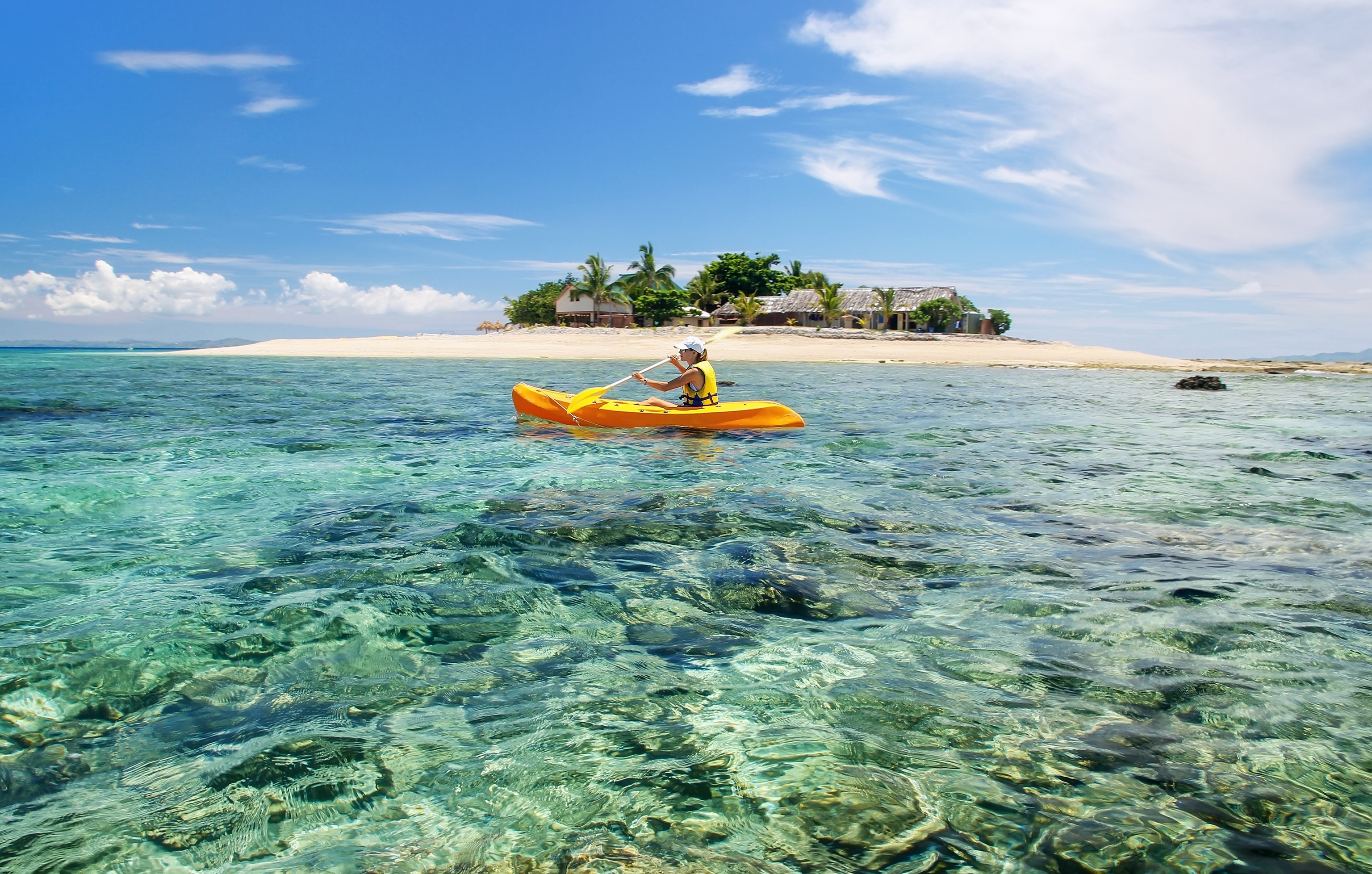 A white woman wearing a life jacket kayaking in clear, aqua sea water with a small tropical island in the background.