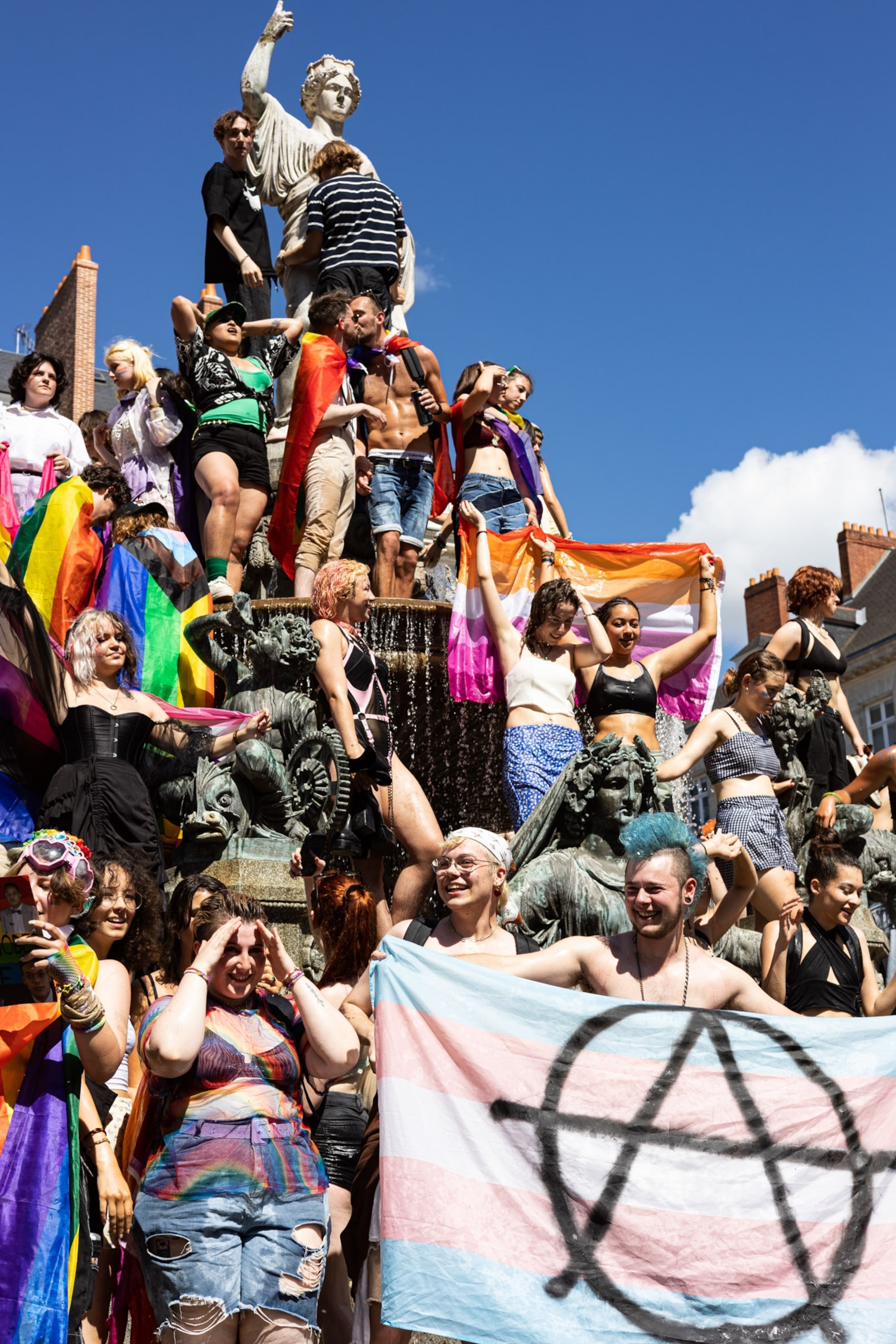 People standing on a statue with a variety of of Pride flags.