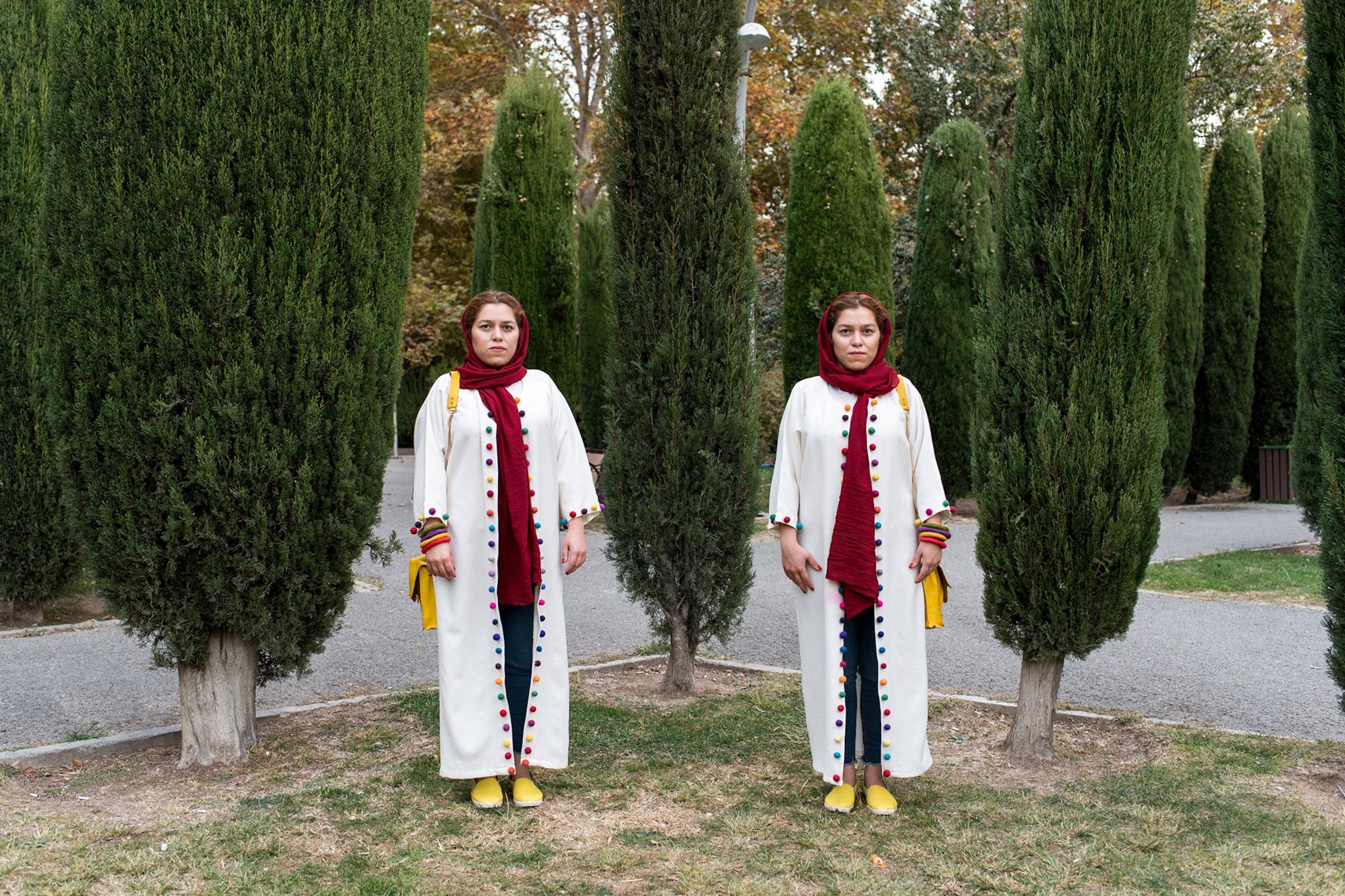 Twins with identical outfits stand near each other with tress behind them.