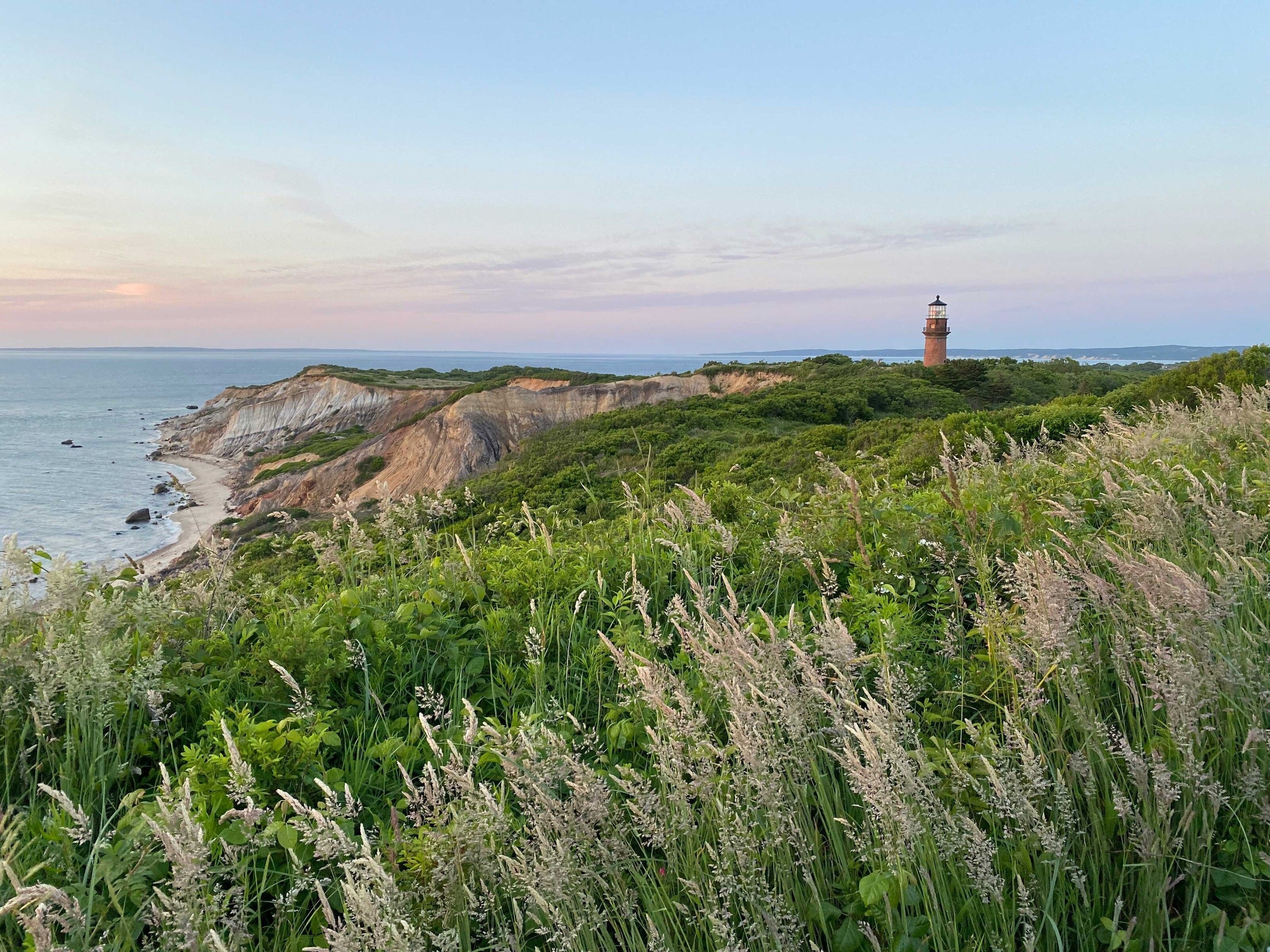 Cliffs and the sea. A lighthouse protrudes from green fields in the distance.