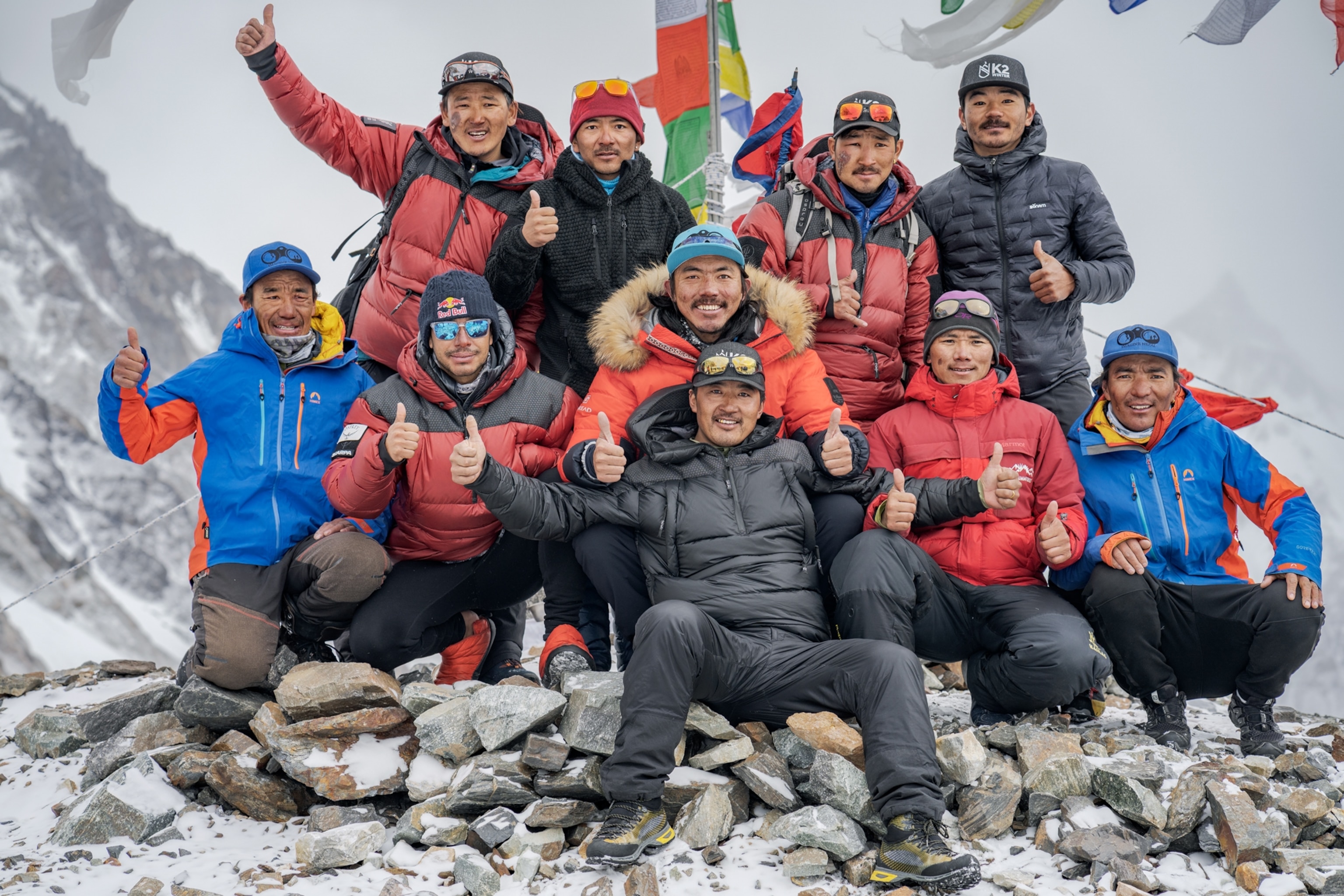 Picture of ten men posing for photograph with their thumbs up under national flag.