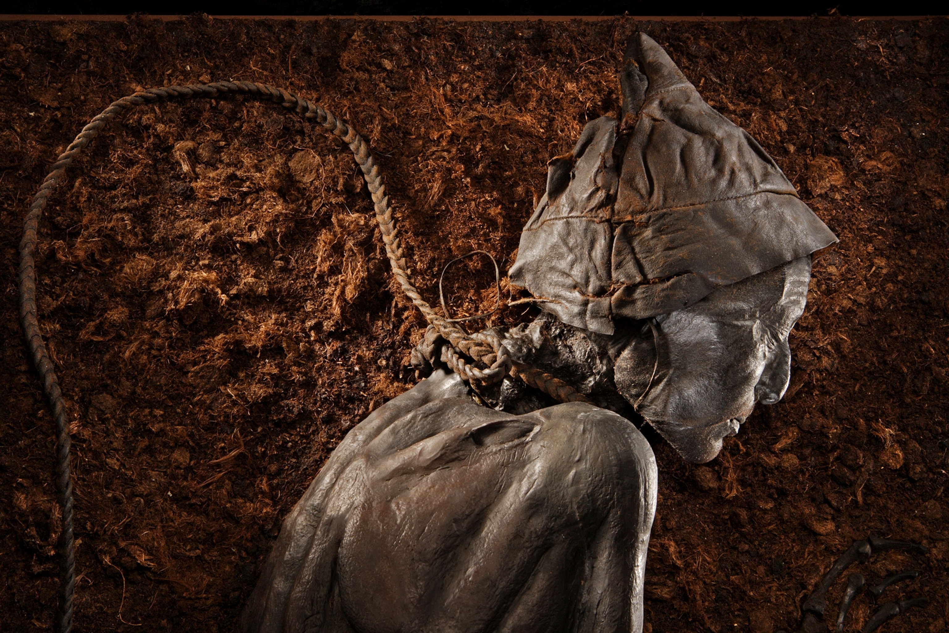 A profile view of an old man from the mid-back and up. The skin appears wrinkly and is colored brown and bronze-like. There is a cap on the head, that is also wrinkly and colored brown and bronze-like. There is a braided rope around the neck and extends in a loop behind him.