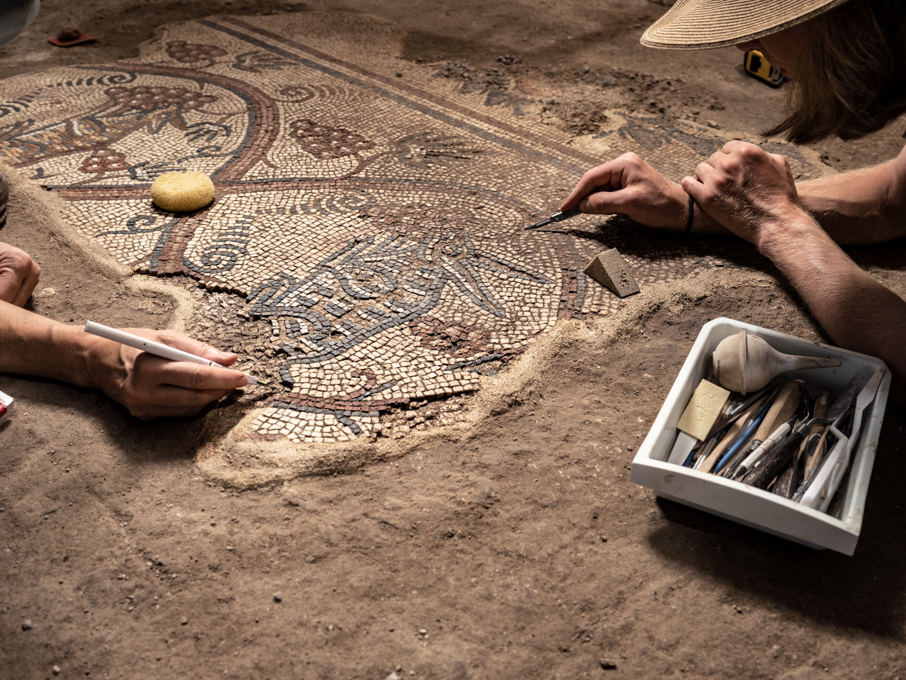 Two persons working with sharp tools on floor mosaic fragment.