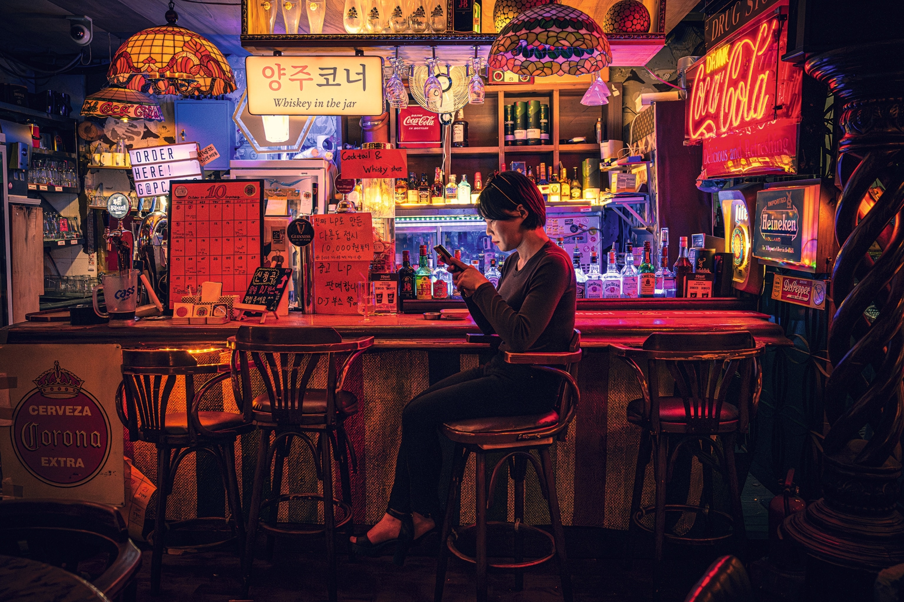 A patron in a bar in Seoul is on their phone. There are neon signs and lamps throughout the bar, with no other person in sight.
