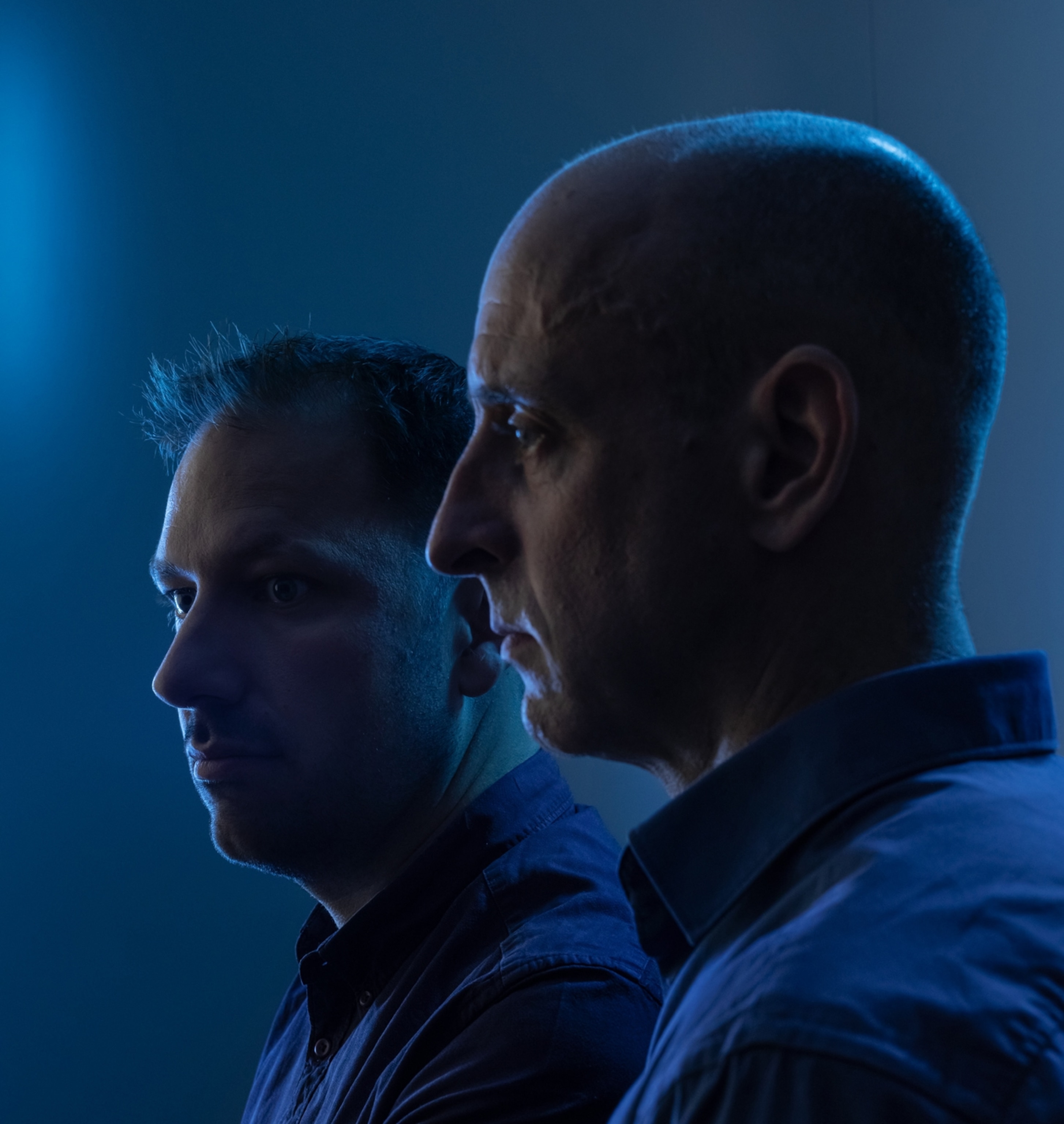 A profile image of two men; they are standing near each other. The lighting is heavily blue with a slight glow around their faces.