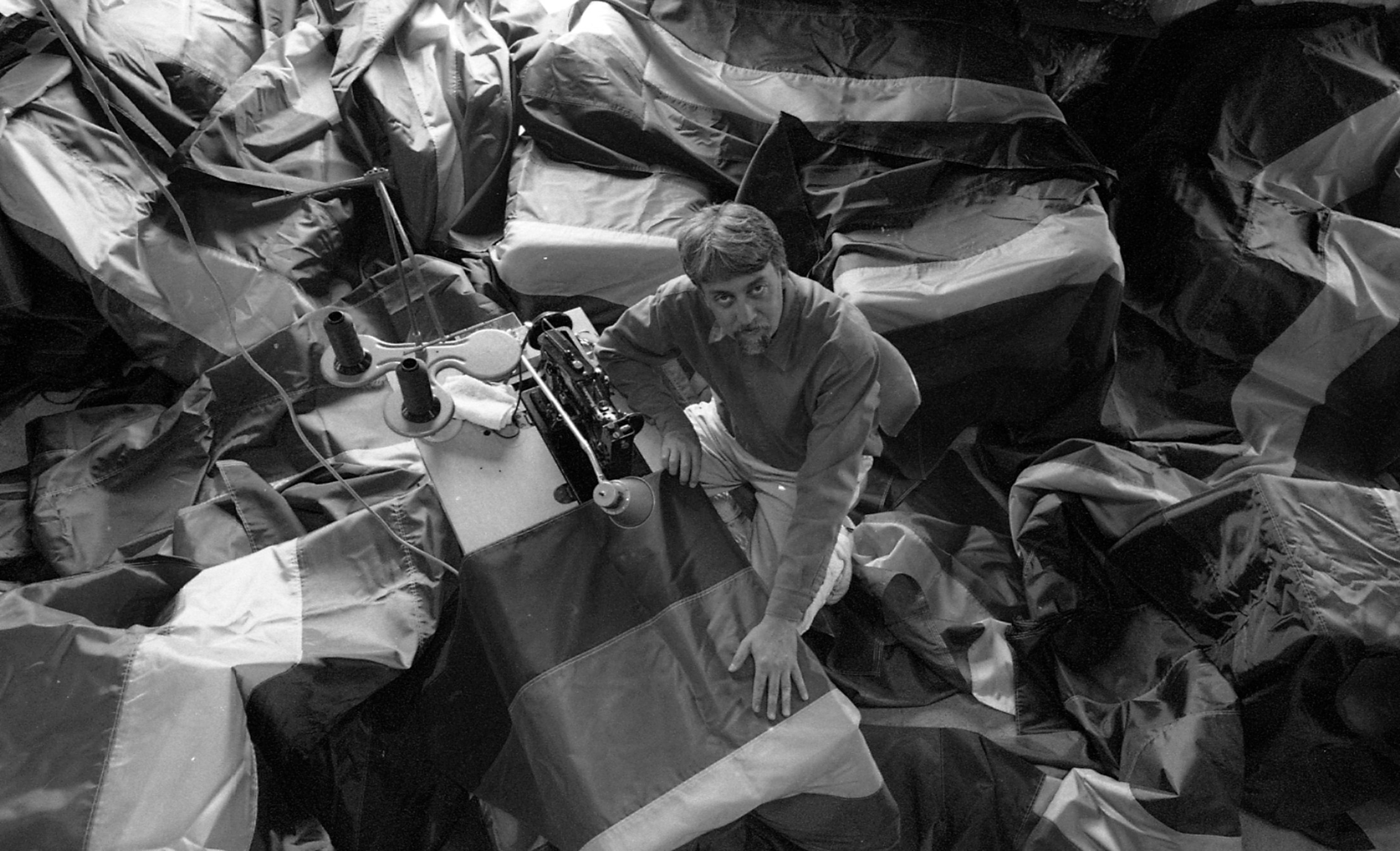 A top view of Gilbert Baker in black and white the surrounded by flags at a sewing machine.