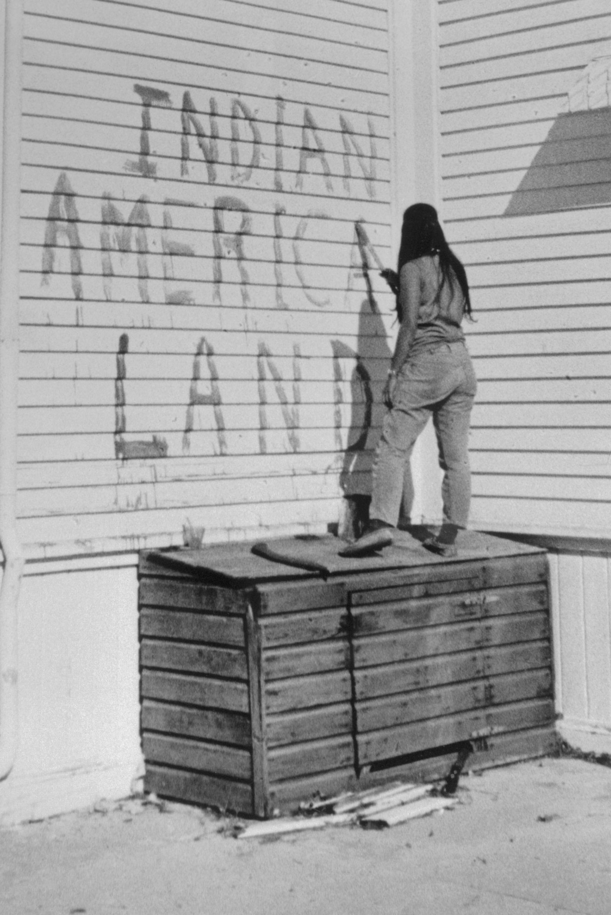 A person stands on a wooden shed and writes Indian America Land on a wall