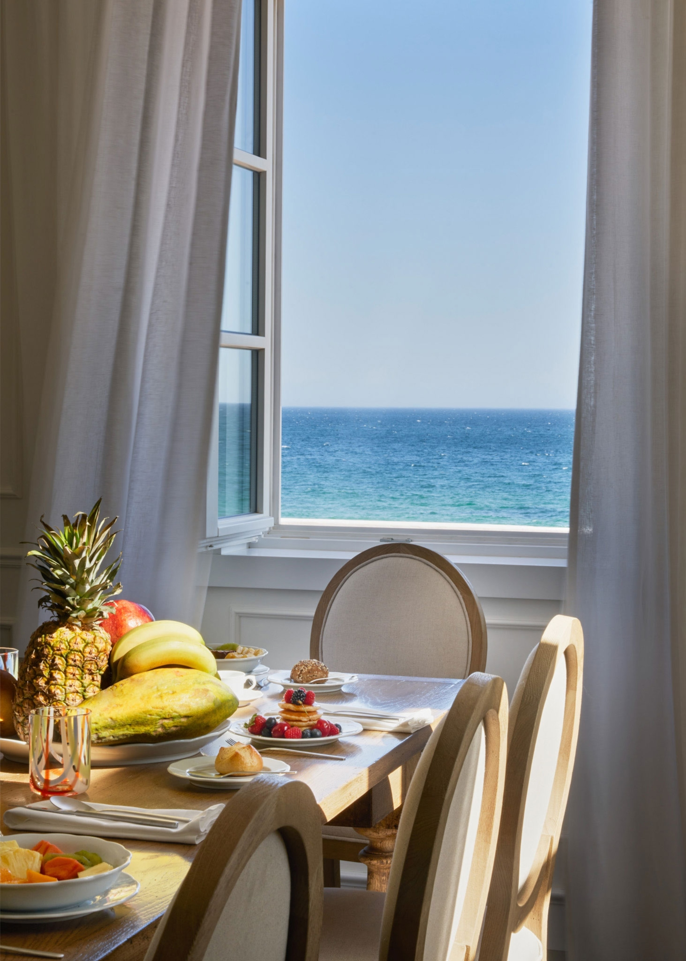 A table full of fresh fruit sits in front of a window that looks out to a blue ocean.