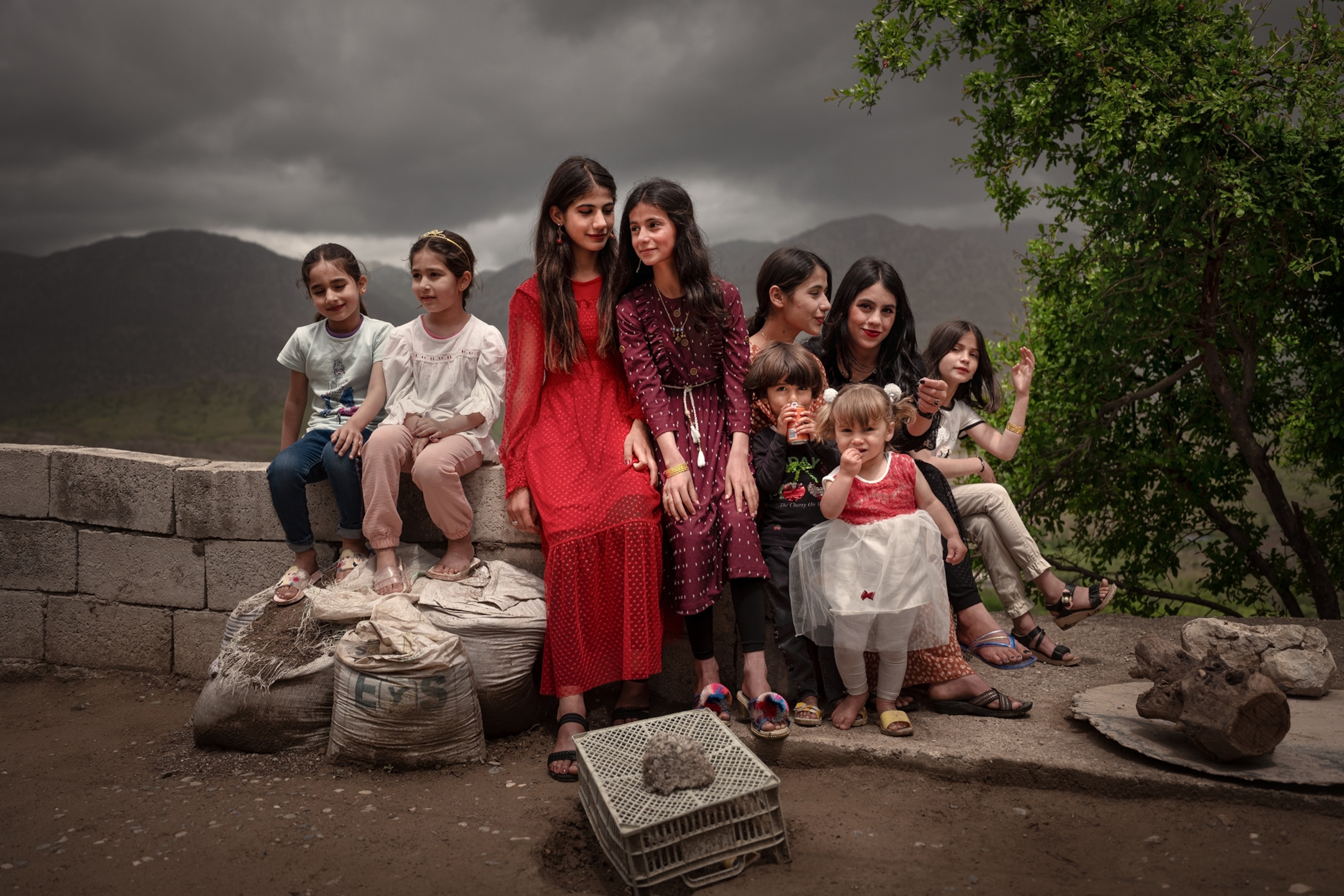 A group of nine young cousins sit playfully along a gray cinder block wall underneath a stormy grey sky.