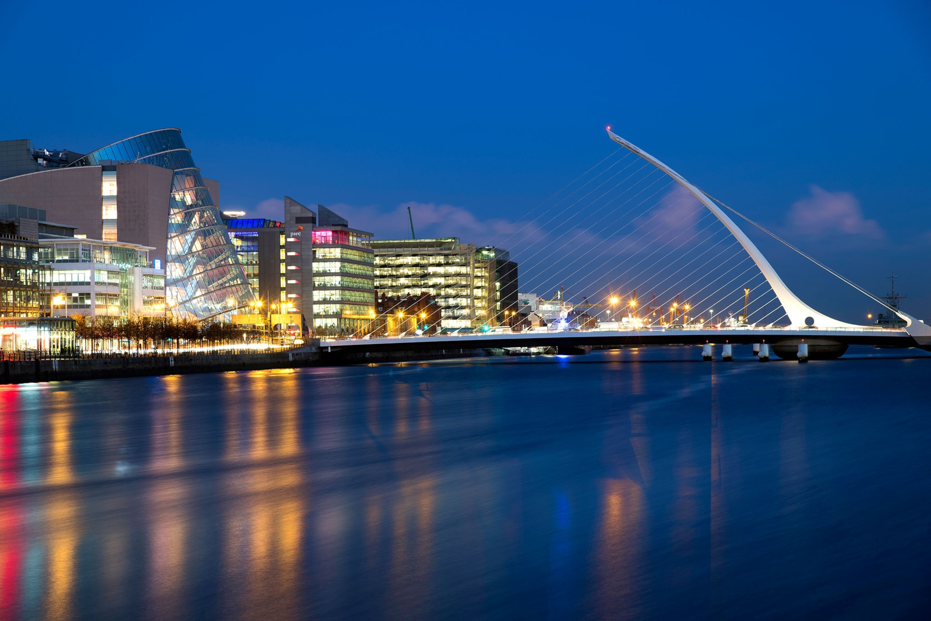 a smooth river reflects light from the city to the left, a white, large, fin shaped bridge is in the distance.