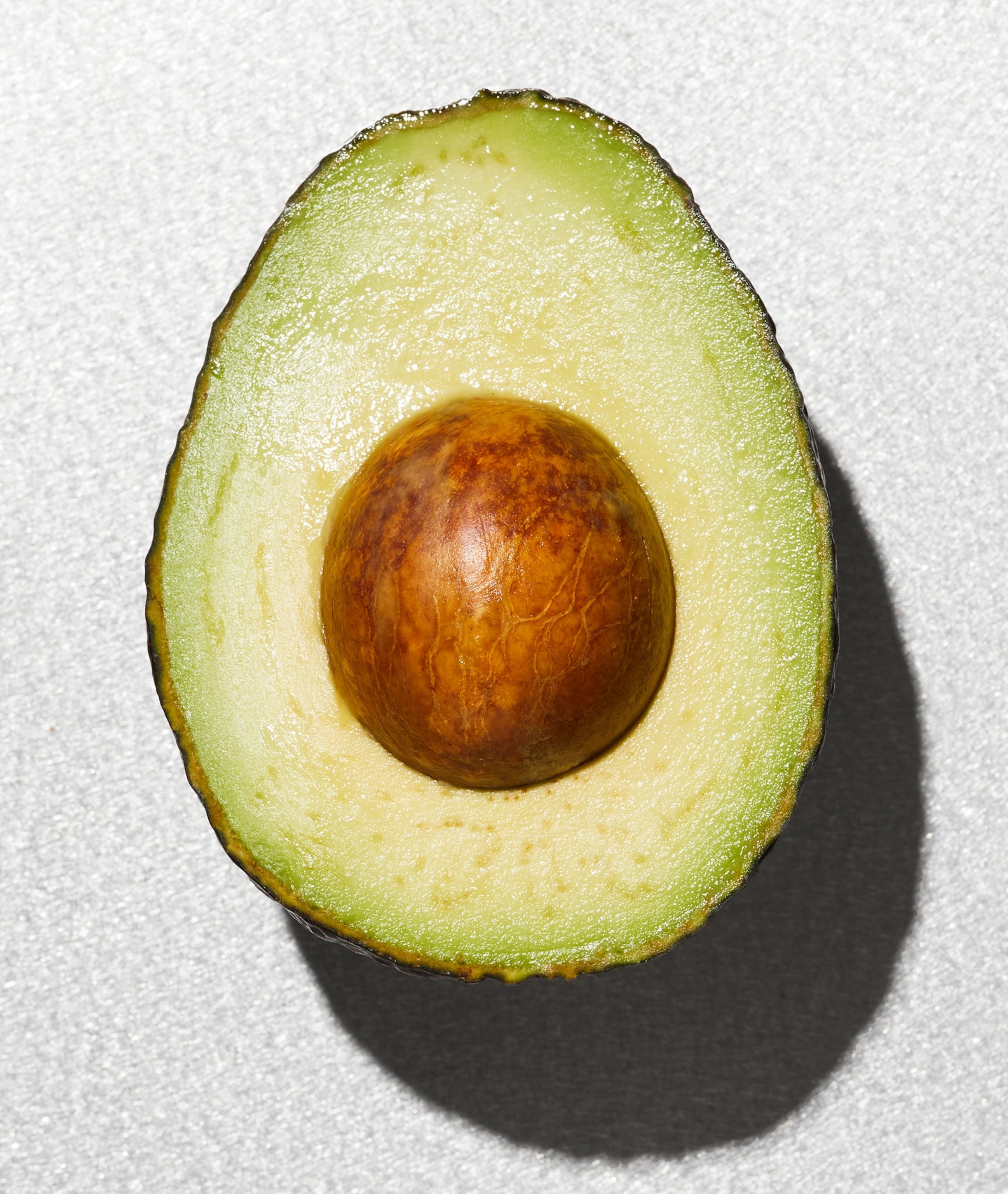 An avocado with dark green skin, a light greenish yellow inside, and a large, round, brown seed in the middle on a white background.