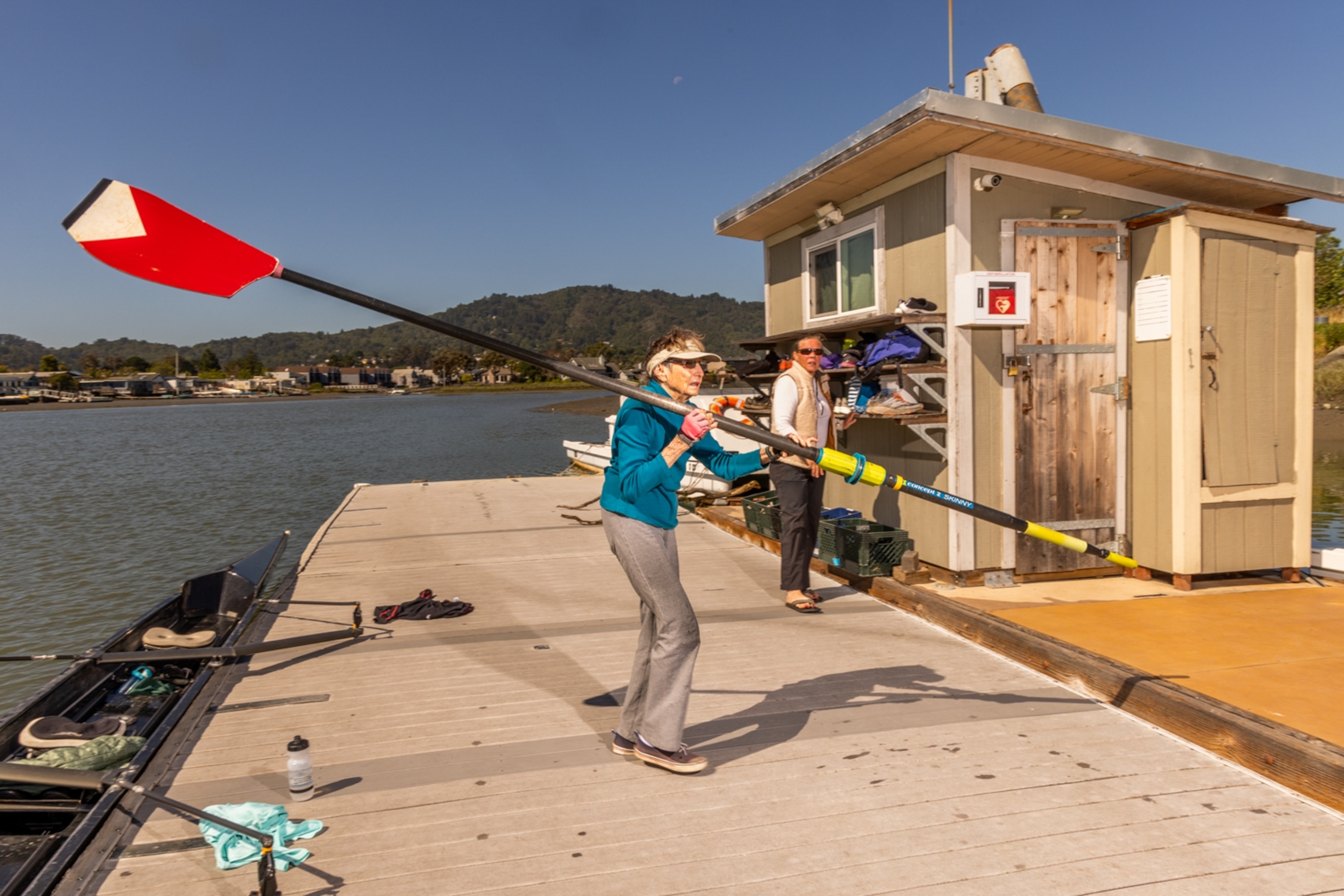 A woman with short grey hair holds an oar double her size under a blue sky.