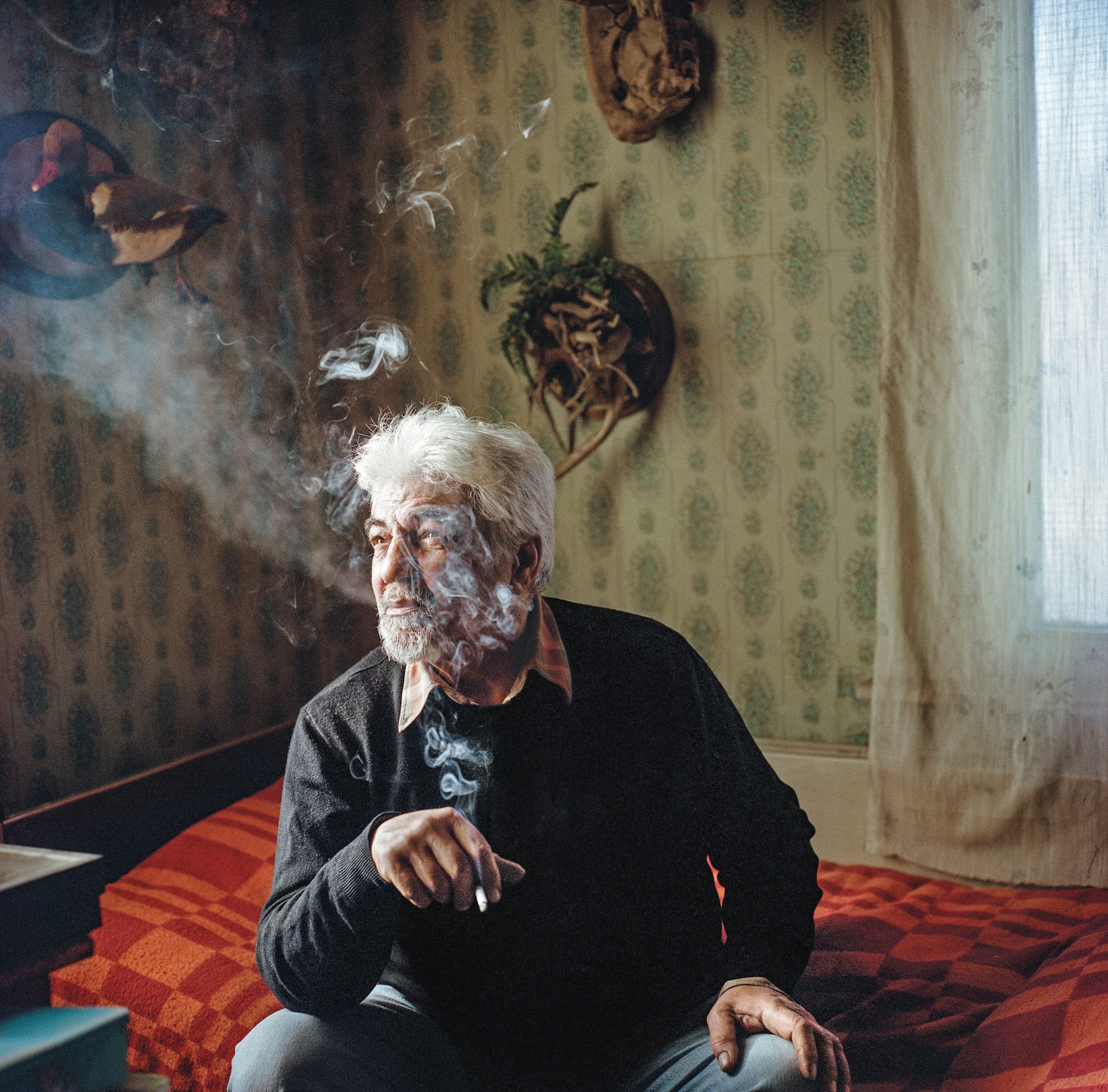 A man sitting on a bed blows smoke while smoking a cigarette.