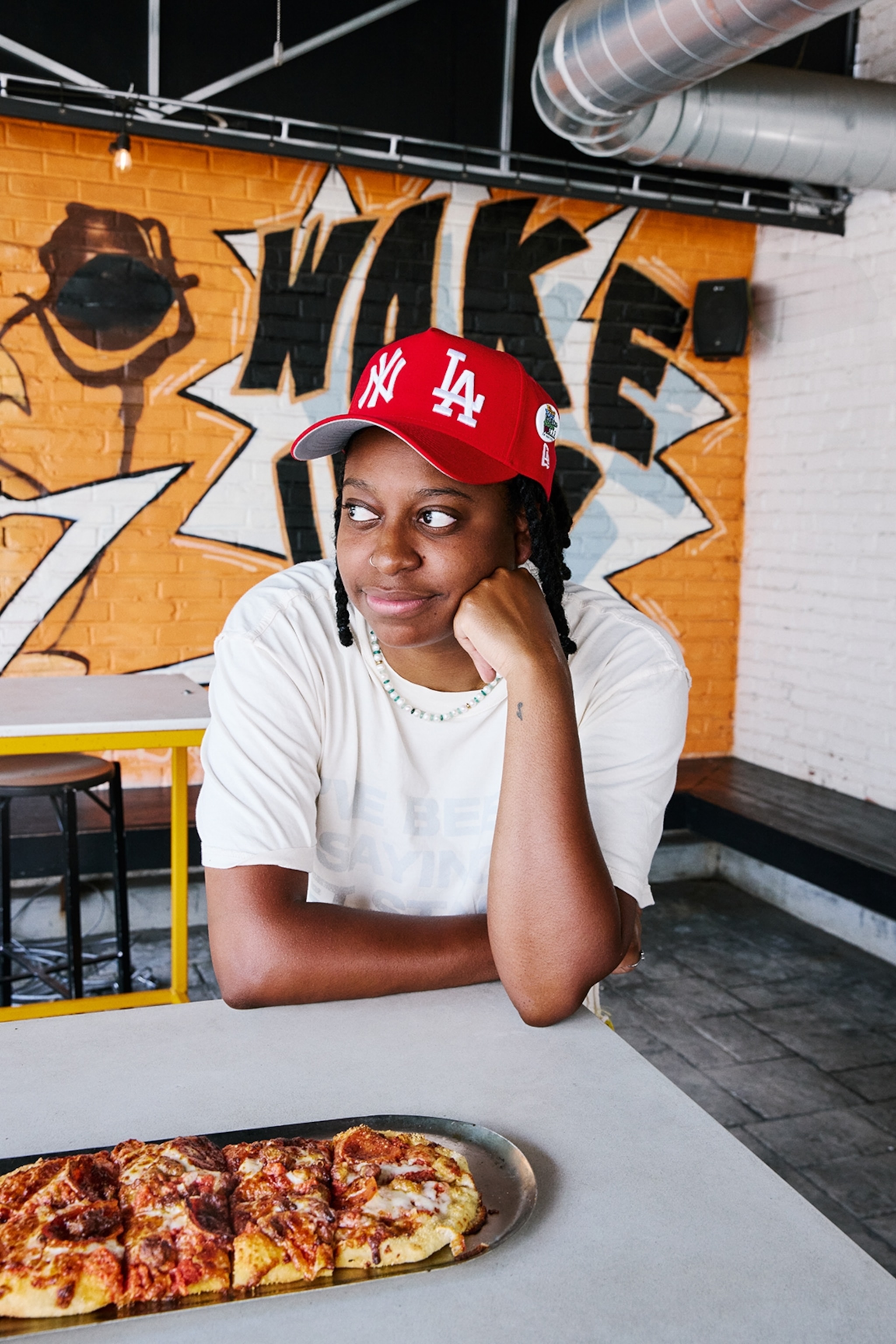 A black woman in her twenties sitting at a tall table and dreamily looking out the window.