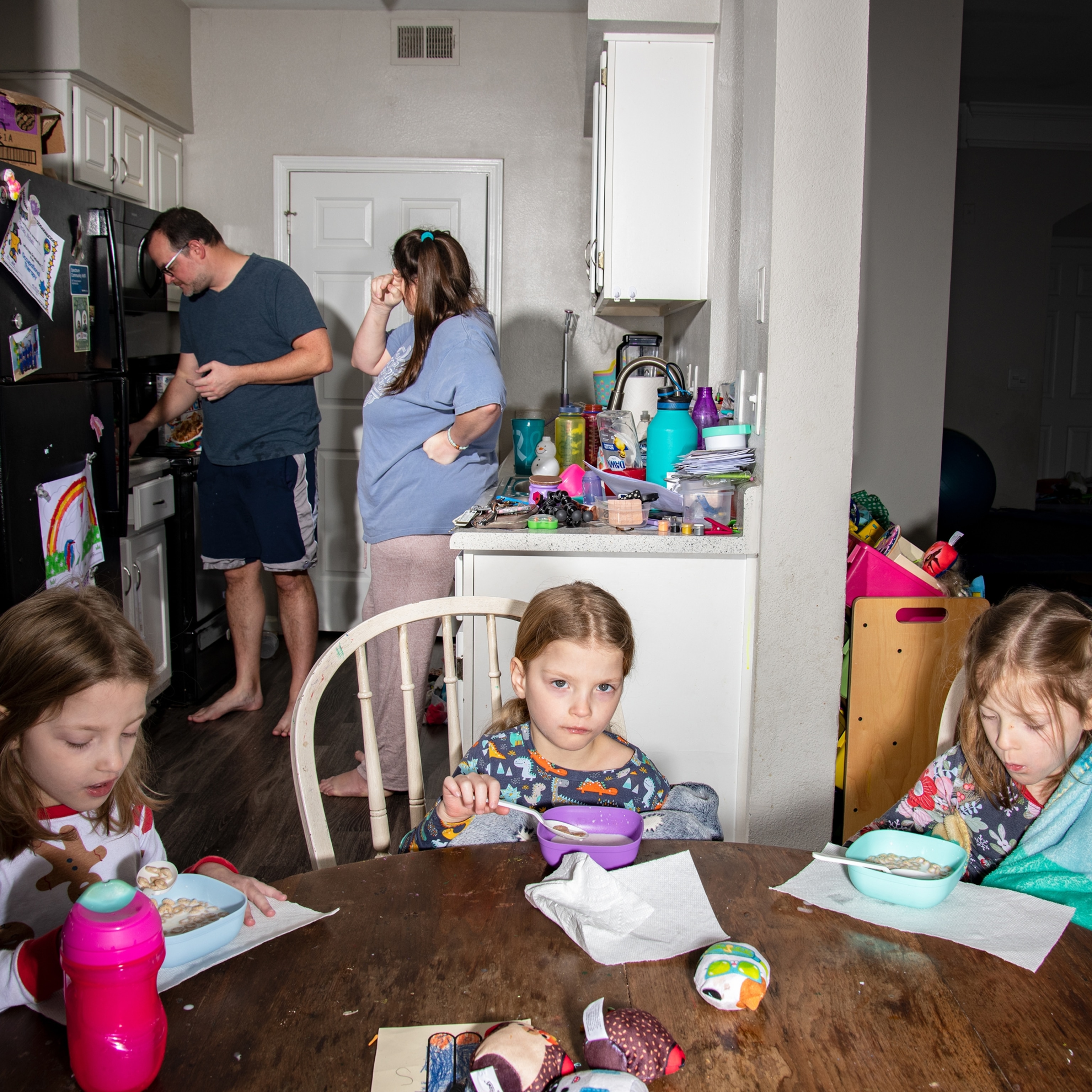Three young girls eat bowls of cereal at the dining table as their mother and father stand distracted in the back of a cluttered kitchen.
