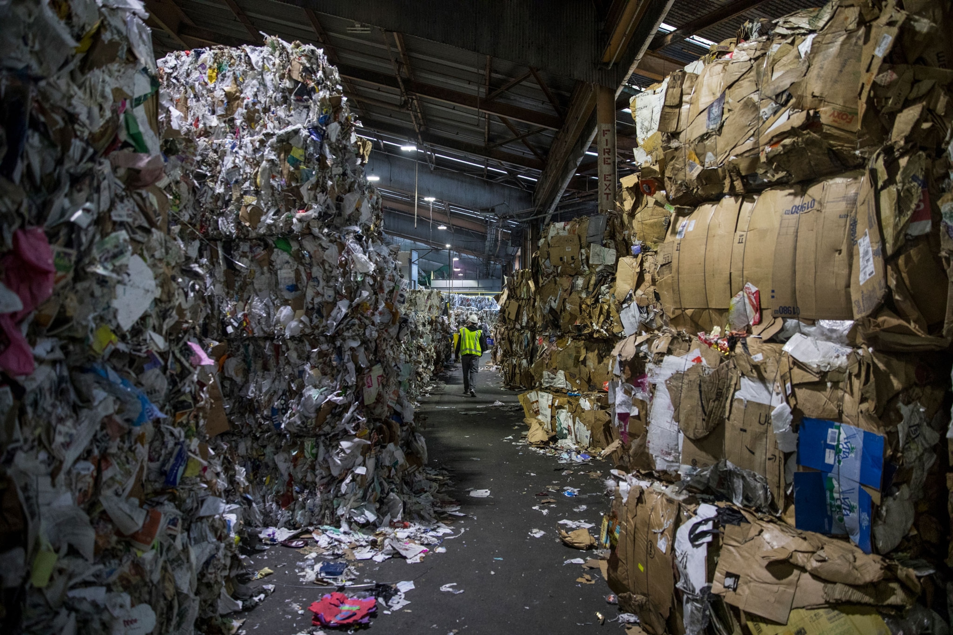 a person in a yellow safety vest walks down a row of piled up recyclable materials towering above their head