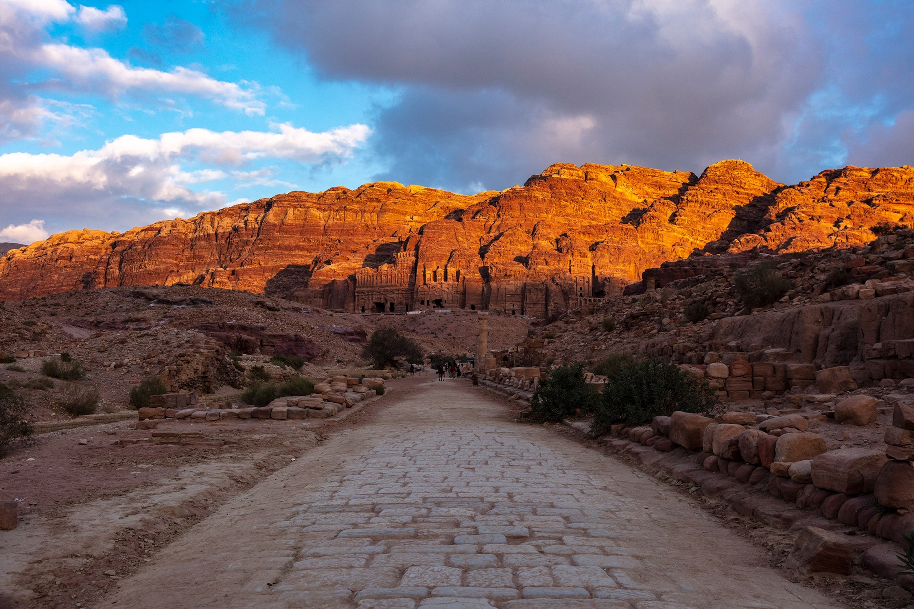 A walk way is in shadow as an old city built into the cliff is illuminated by light.