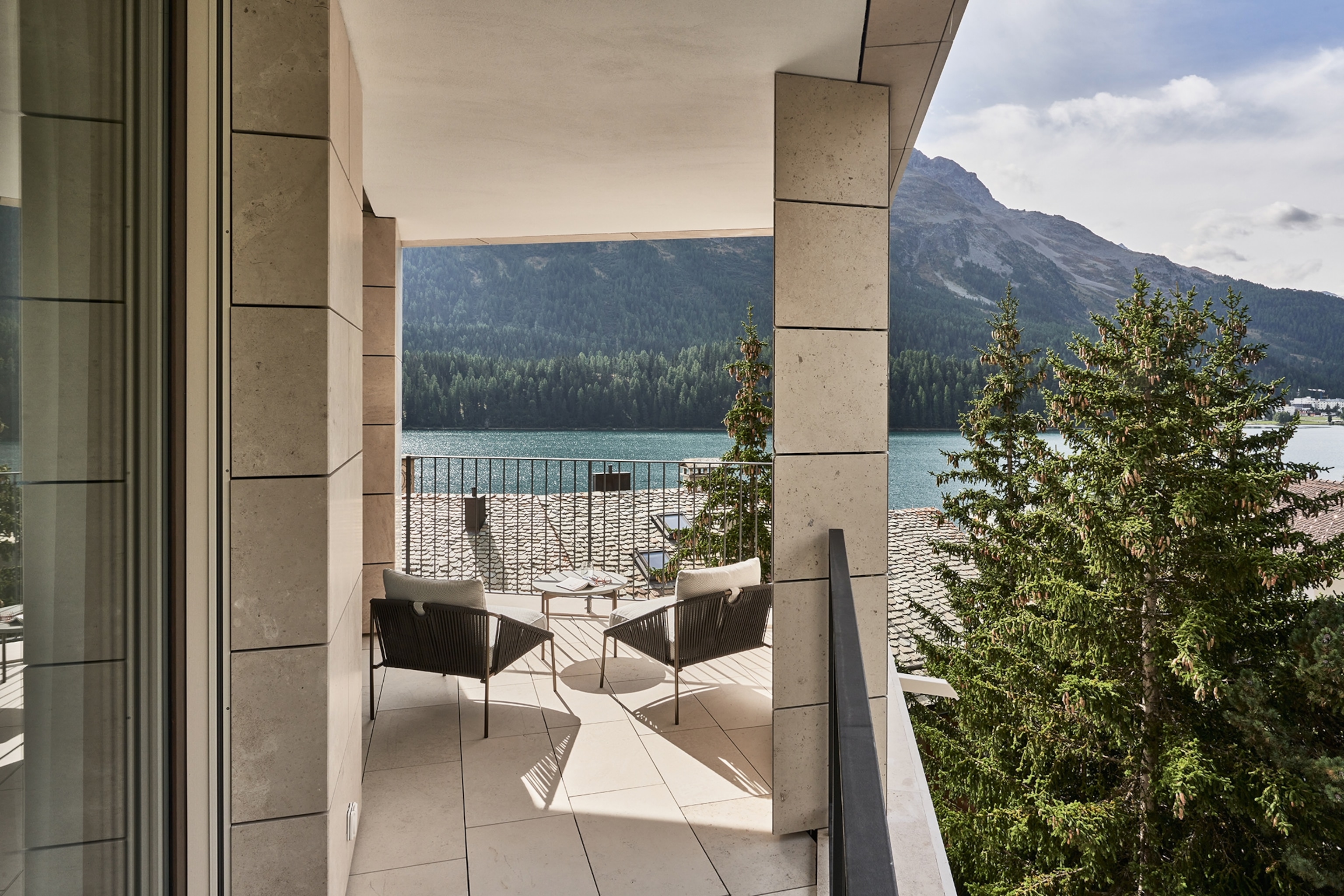 A view of a lake from a balcony in a luxury hotel in St Moritz. Two outdoor chairs and a table face the lake.