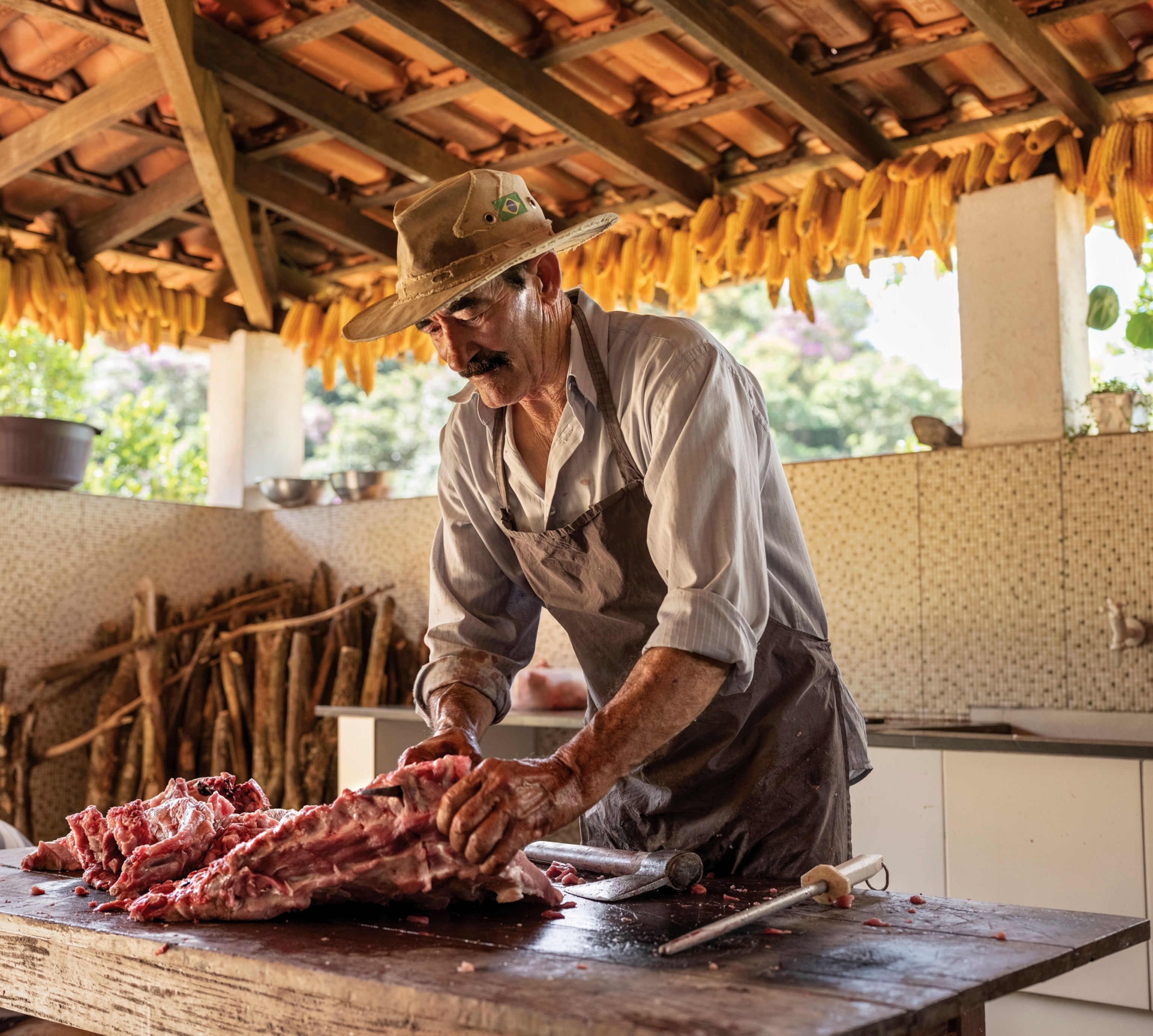 A man wearing a hat in an open air thatched roof room, cuts into a large slab of meat.